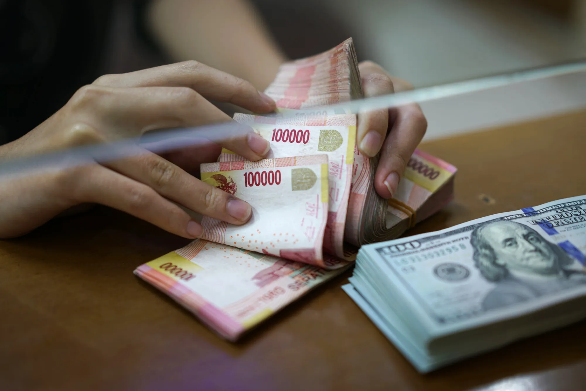 An employee counts Indonesian 100,000 rupiah banknotes as a bundle of U.S. one-hundred dollar banknotes sits on a counter at a currency exchange store in Jakarta, Indonesia, on Saturday, Aug. 11, 2018.&nbsp;