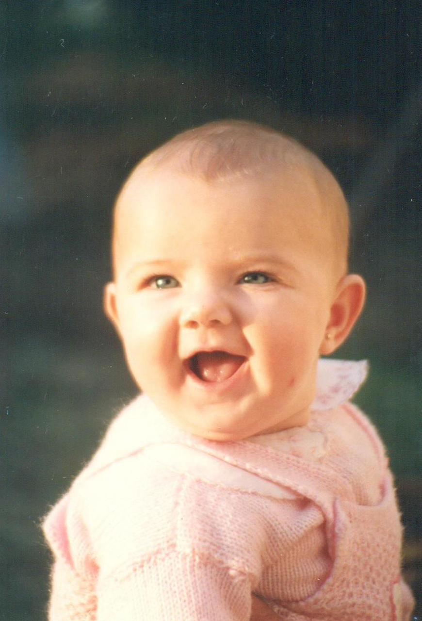 A photo of a caucasian smiling baby wearing a pink knit ensemble against a blue background.