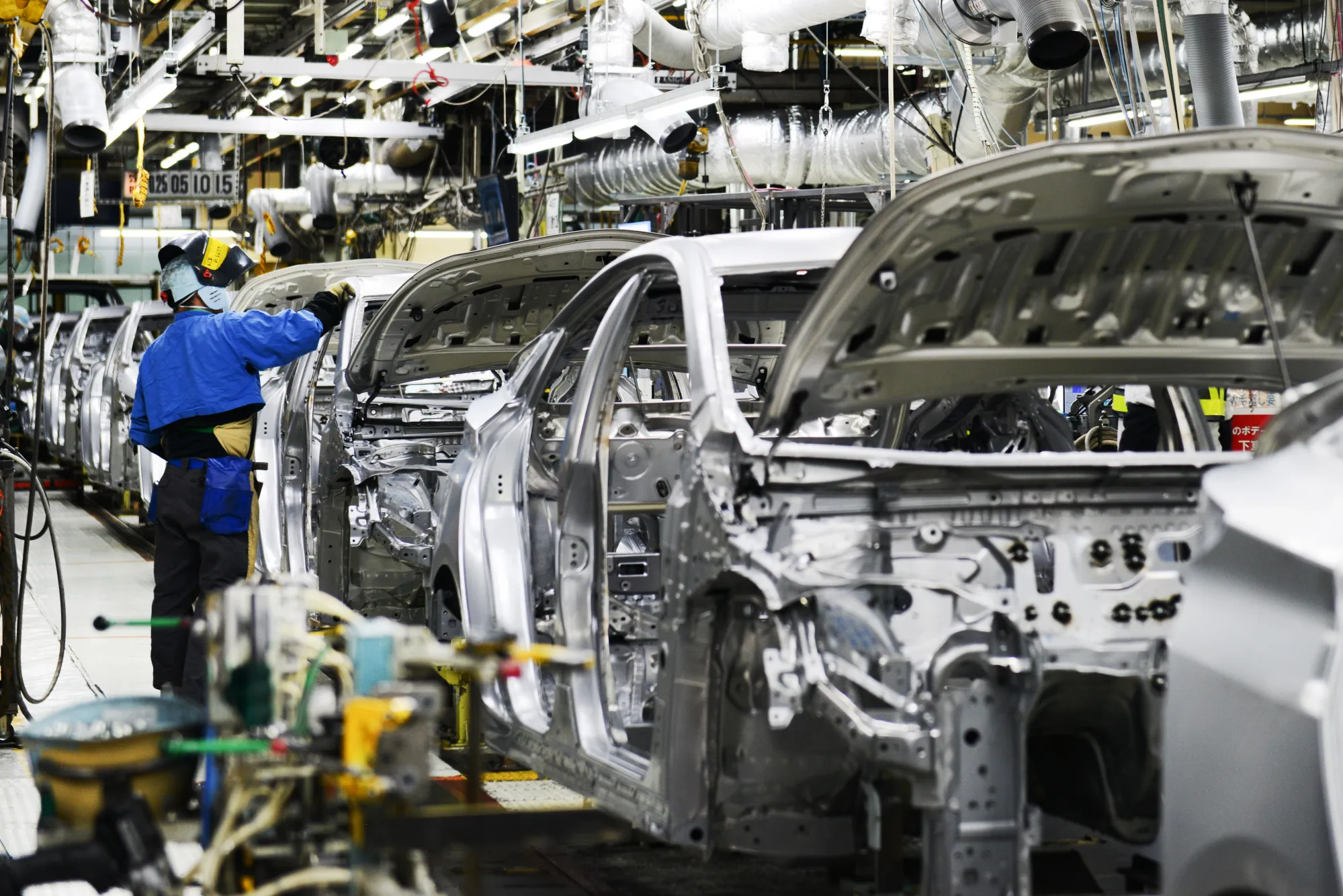 A worker stands next to vehicles moving along a conveyor on the Prius hybrid and Priyus plug-in hybrid vehicle (PHV) production line of the Toyota Motor Corp. Tsutsumi plant in Toyota City, Aichi, Japan, on Saturday, Dec. 8, 2017.&nbsp;