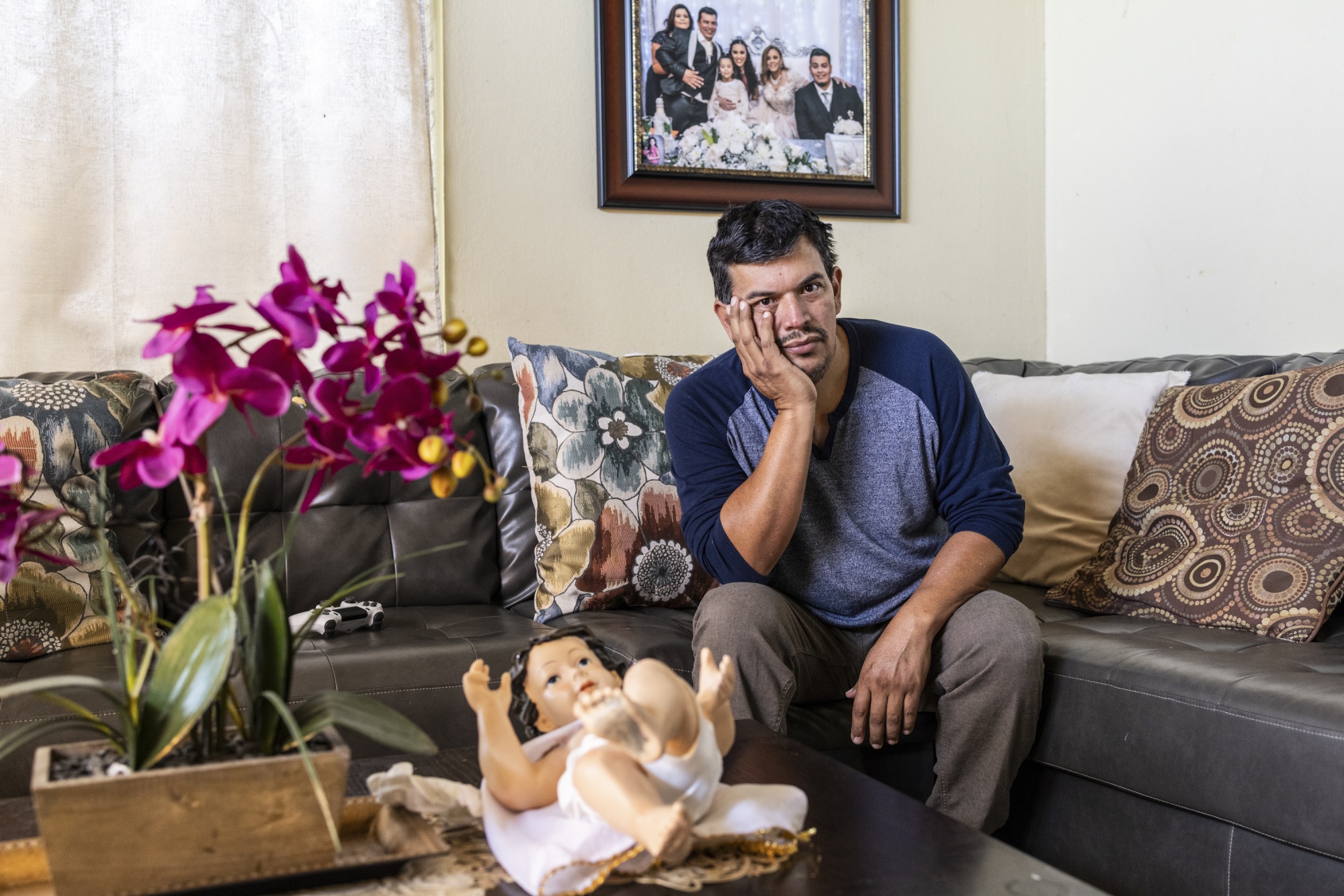 A man sitting on a couch, resting his head on his hand looking pensive, next to him lies a game remote control and above him is a framed image of his family.