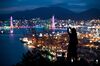 A woman uses a smartphone to take a photograph of the illuminated Port of Busan in Busan, South Korea, on Sunday, July 16, 2017. South Korea's exports will continue to rise in July and the third quarter as global trade continues to recover and unit costs rise, especially in sectors including semiconductors, vessels, petroleum goods and steel, according to a statement from the trade ministry.