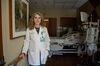 Doctor Shelley Stanko stands against the wall of a hospital room, holding a handrail, in front of an unoccupied hospital bed.