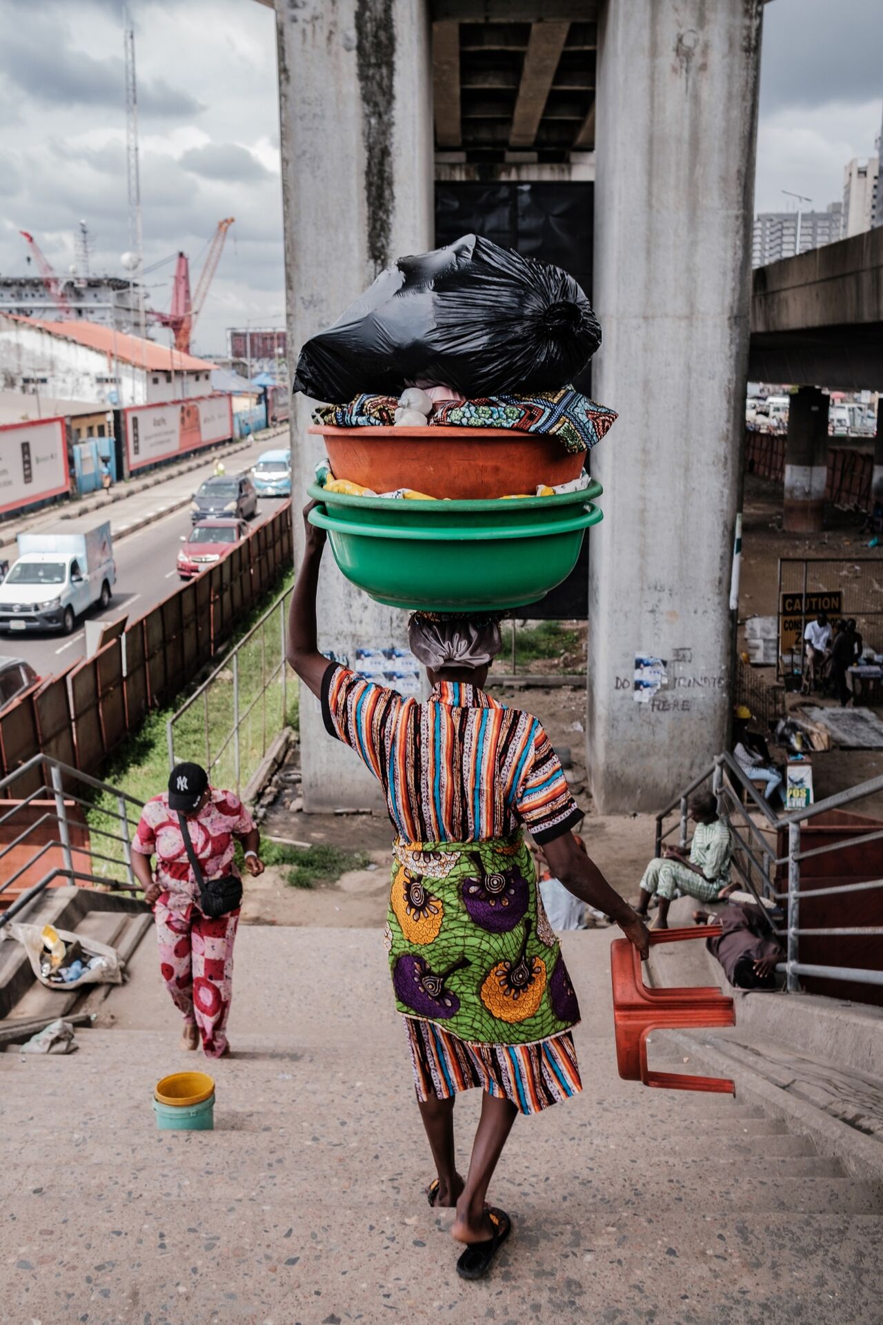 A street vendor in Lagos, Nigeria, walking on a pedestrian bridge carrying stacked bowls and goods on her head beneath an overpass.