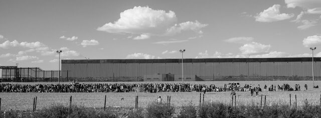 A crowd of asylum seekers at the US-Mexico border wall wait to be processed by US Customs and Border Protection agents in TKTK. 