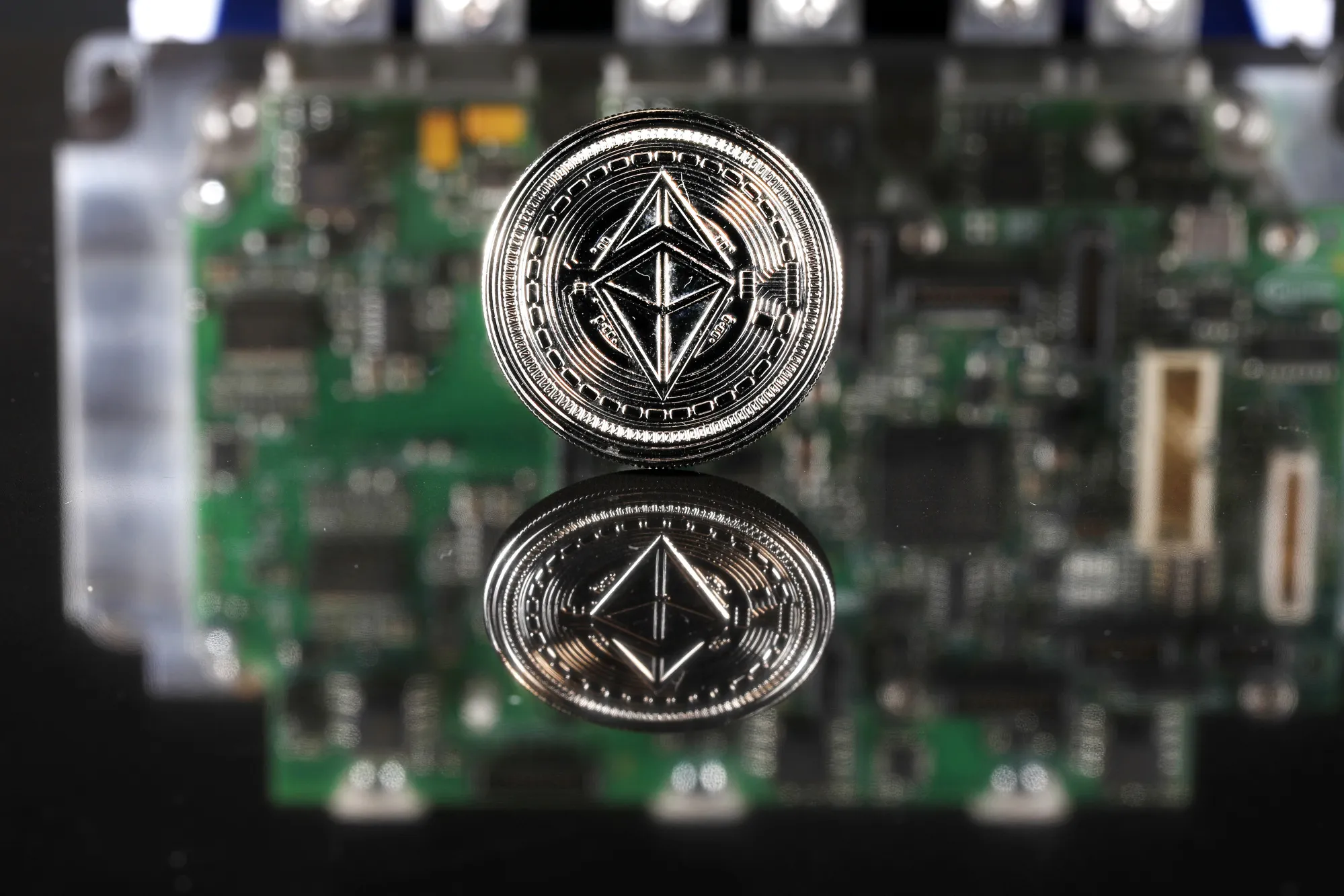 A coin representing Ethereum cryptocurrency sits reflected on a polished surface and photographed against a computer circuit board.