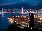 A woman uses a smartphone to take a photograph of the illuminated Port of Busan in Busan, South Korea.