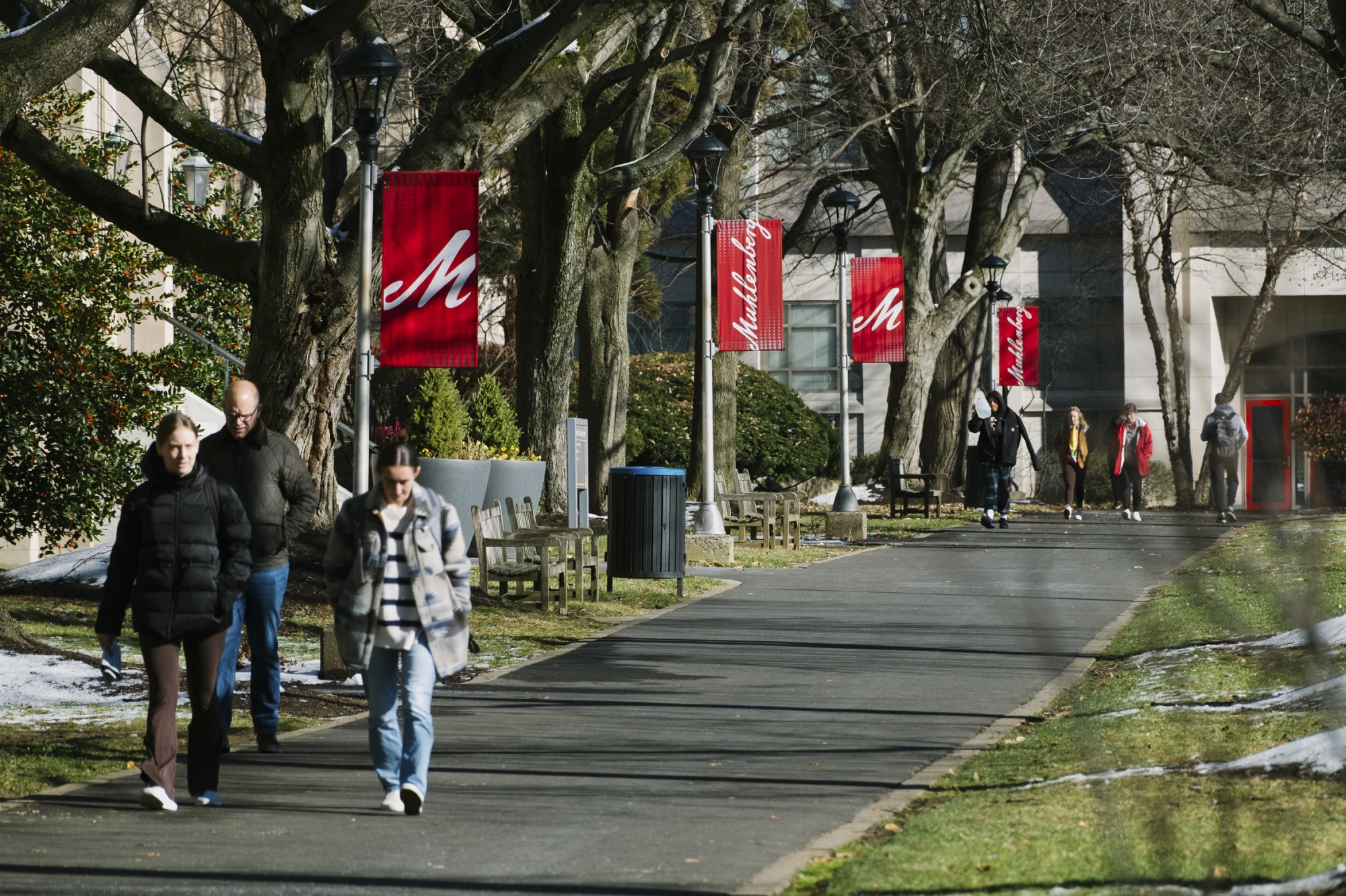 学生们走在大学校园里。地面上可见斑驳的积雪，学生们都穿着冬装。校园的标志在旗杆上清晰可见，一个醒目的白色字母“M”印在红色背景上（代表穆伦伯格学院）。