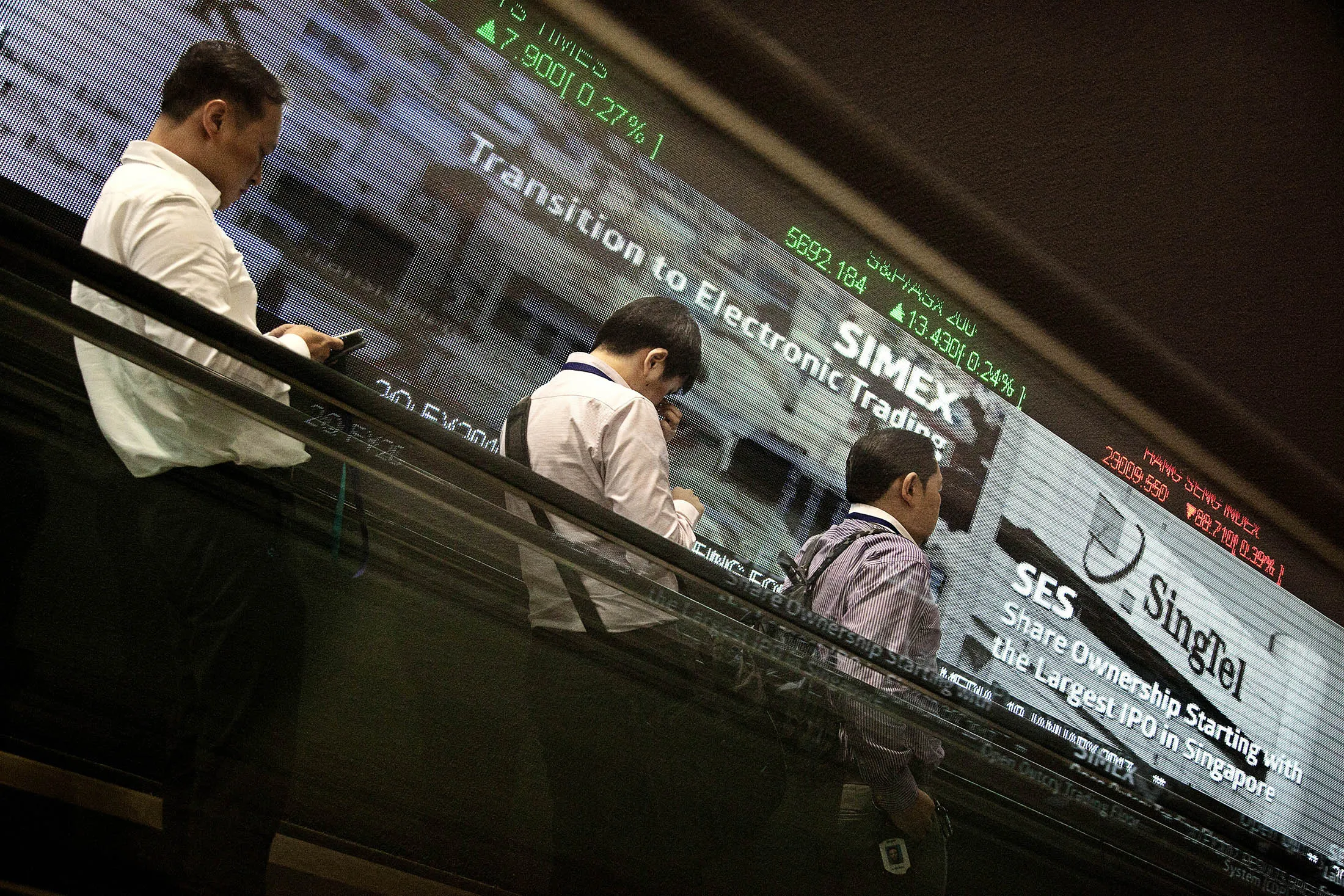 Men ride on an escalator past an electronic screen and ticker board displaying stock figures at the Singapore Exchange Ltd. (SGX) headquarters in Singapore.
