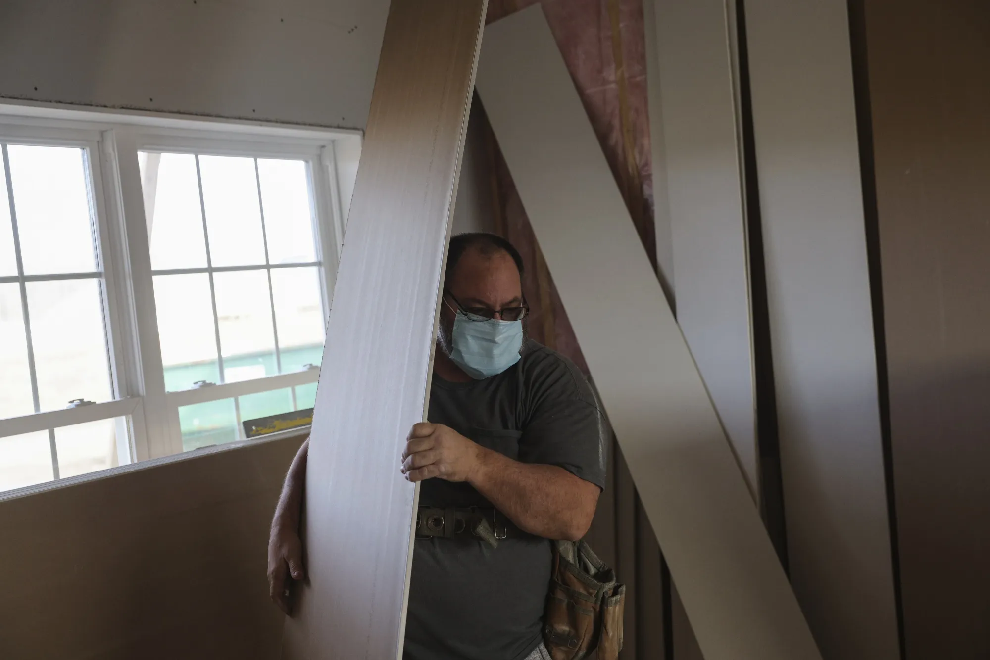 A contractor wearing a protective mask moves pieces of sheet rock while working at a home under construction.