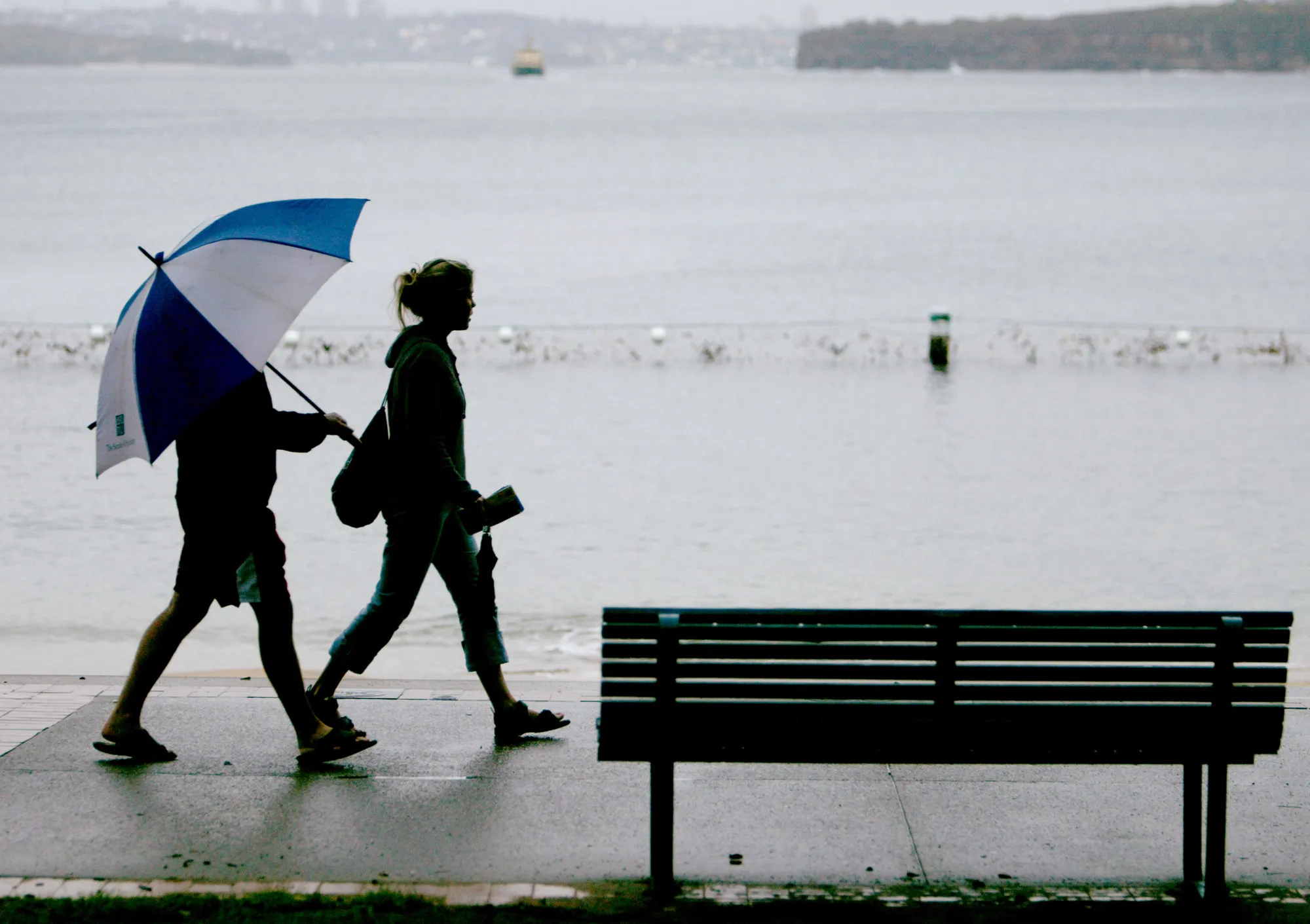 A couple walk through the rain along West Esplanade past an empty seat overlooking Sydney Harbour in the suburb of Manly on the city's North Shore Wednesday, May 14, 2003.
