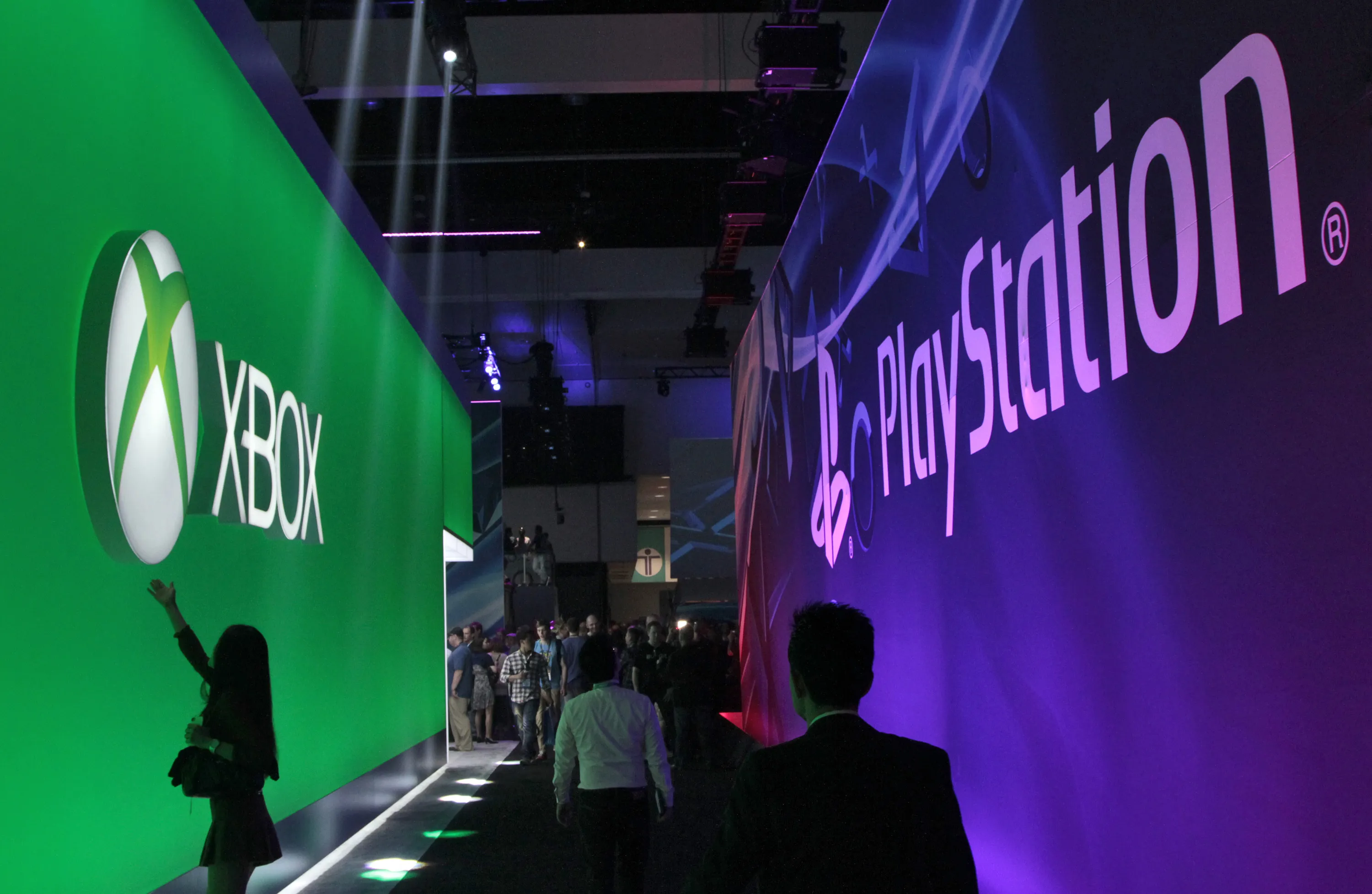 Attendees walk past Microsoft Corp. Xbox, left, and Sony Corp. Playstation logos as a woman gestures at the Xbox logo during E3 2013 in Los Angeles. Photographer: Jonathan Alcorn/Bloomberg