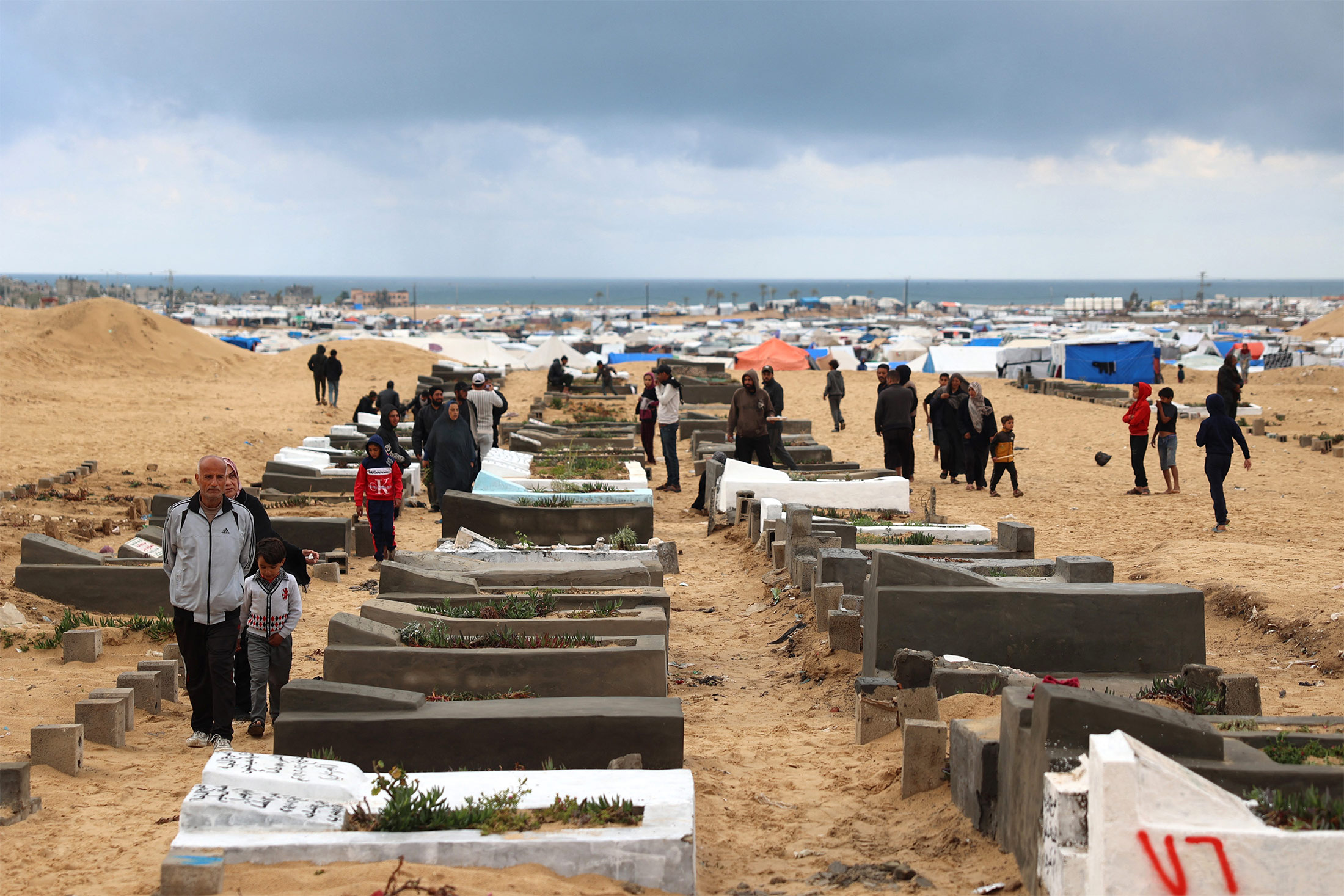 People walk along coffins set up in rows. A large camp of tents and the ocean is visible in the background.