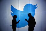 People&nbsp;check mobile devices whilst standing in front of Twitter Inc.'s logo.