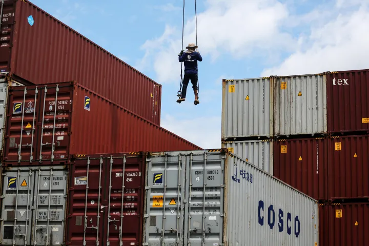 A worker stands on a truck-mounted crane's main hoist line above stacked container.