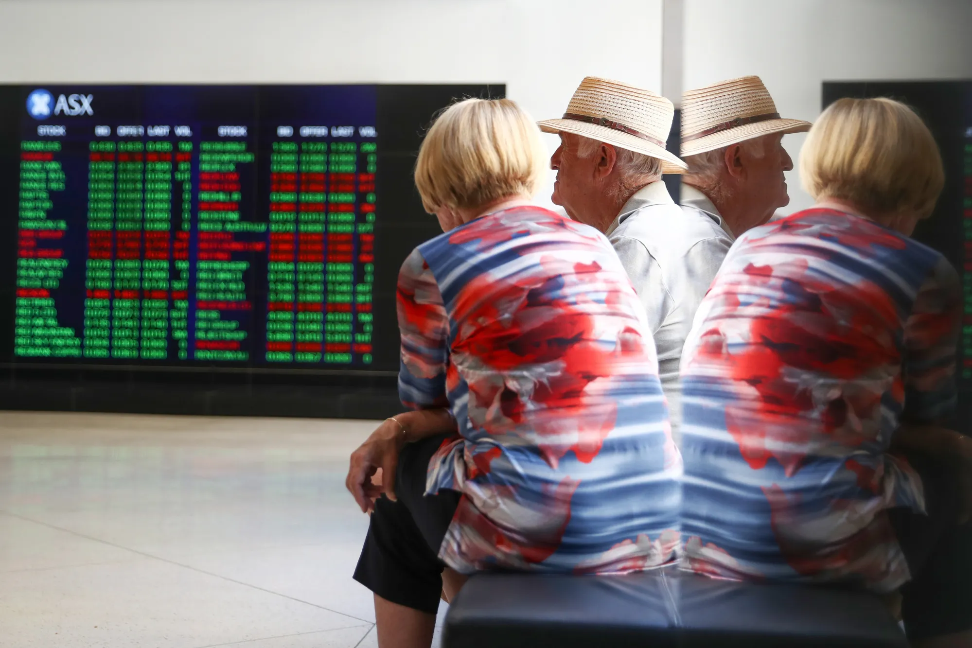 People are reflected in a glass while looking at electronic boards displaying stock information.