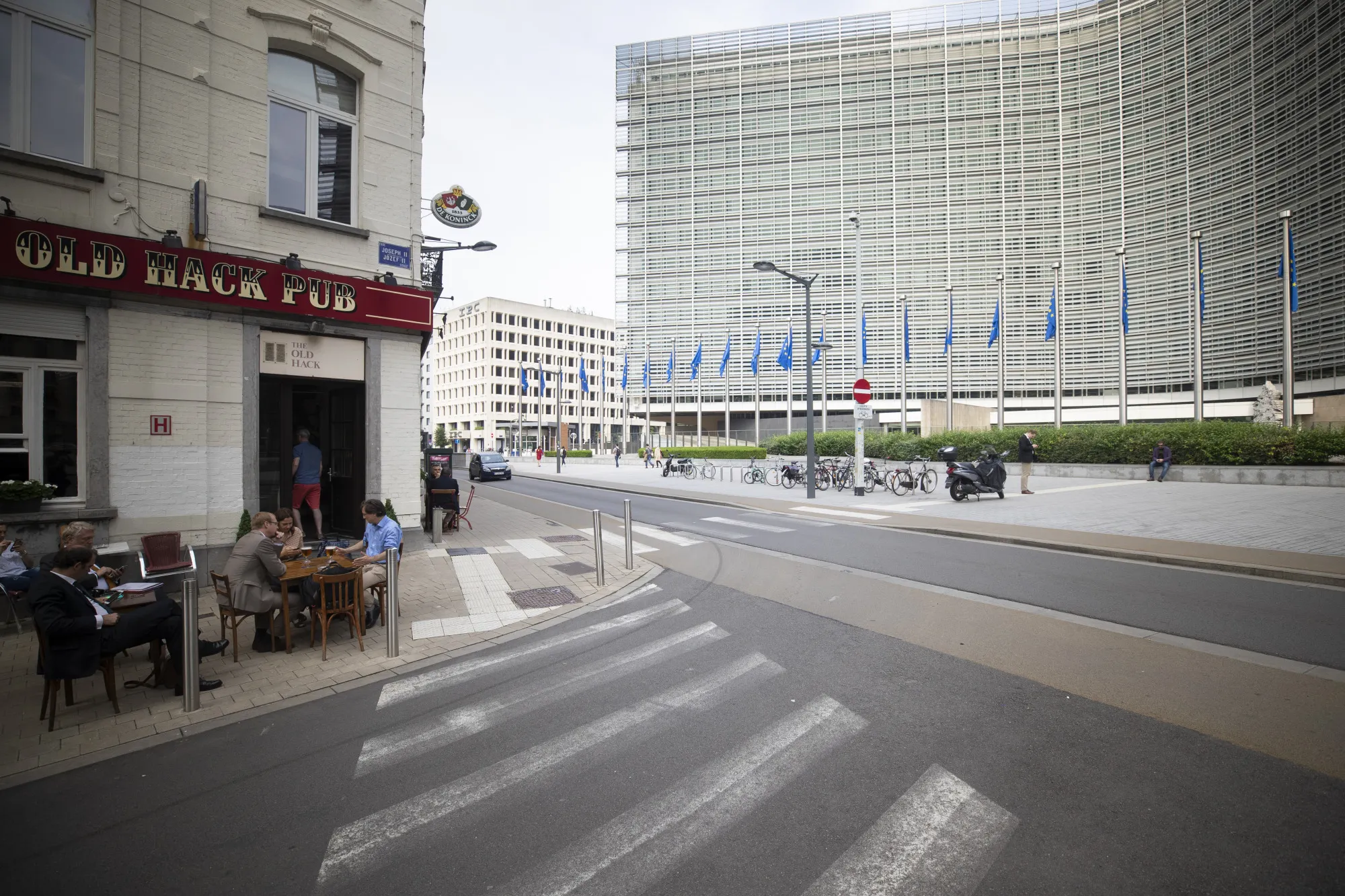 People sit outside the 'Old Hack Pub' in front of the European Commission Berlaymont building as European Union (EU) flags fly in Brussels, Belgium, on Friday, June 8, 2018. Chief negotiator for the European Union Michael Barnier rejected the key elements of U.K. prime minister Theresa May’s plan to overcome one of the thorniest divorce questions, but said talks continue.