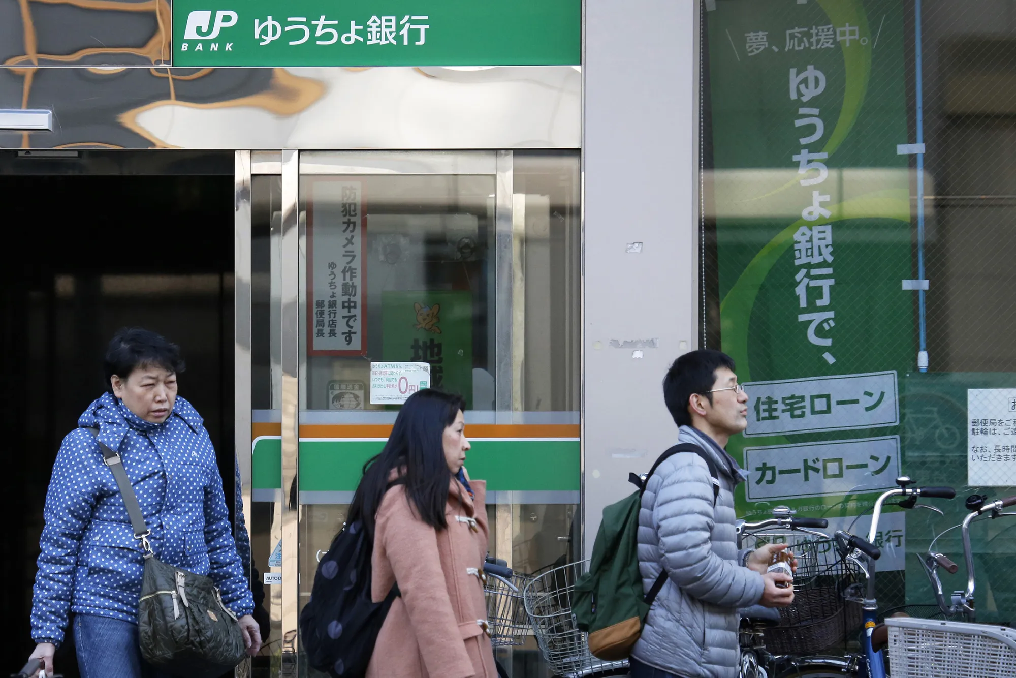 Pedestrians walk past a Japan Post Bank Co. branch in Tokyo, Japan, on Friday, Dec. 26, 2014. Japan's government plans to split Japan Post Holdings Co. into three listed companies by selling shares to the public in August or later as it privatizes the nation's biggest consumer bank.

