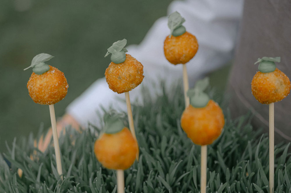 A waiter holding a container of dark green grass stuck with rows of a fried savory canape that looks like a delightful orange lollipop topped with a piped green sauce and finished with a leaf from a herb.