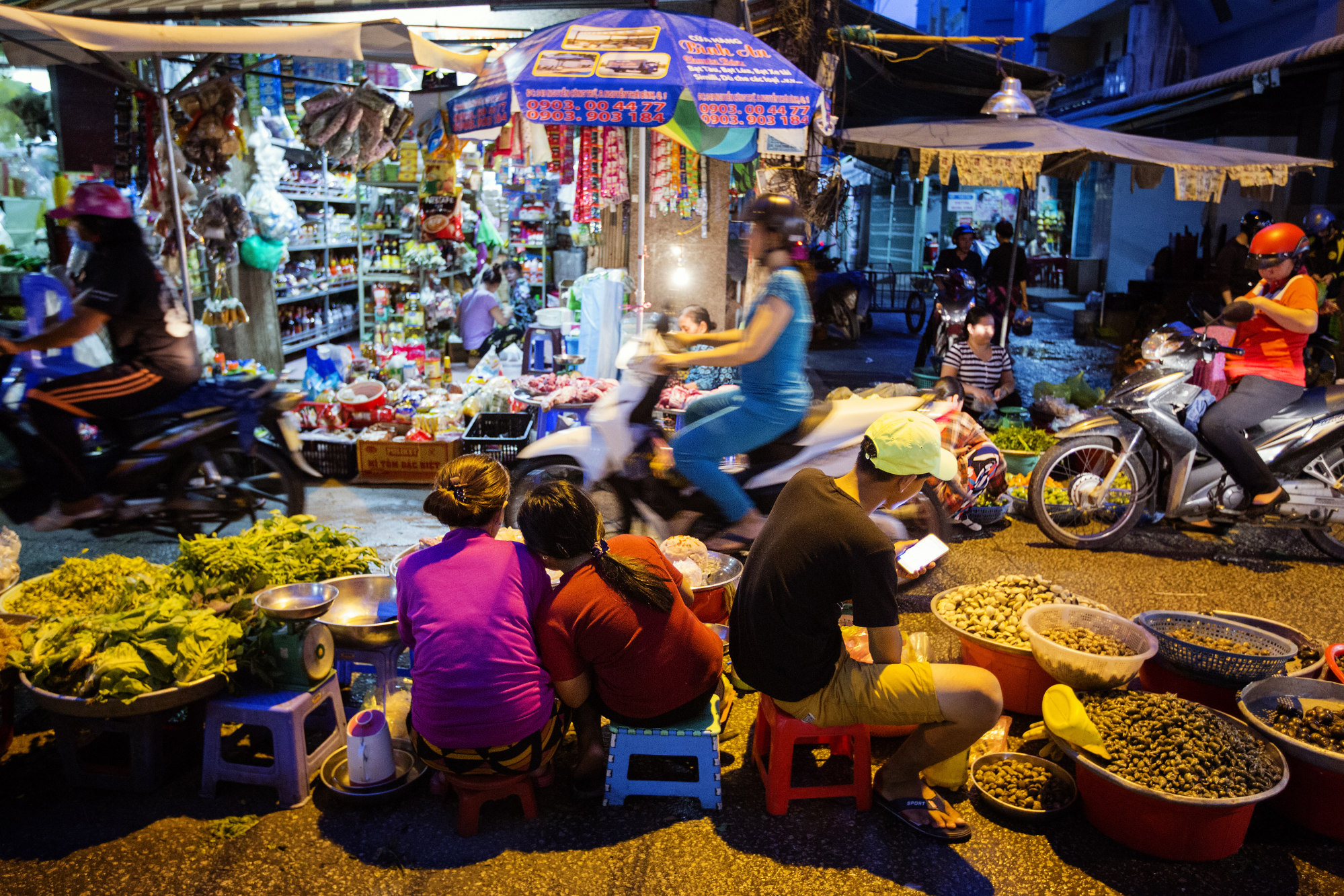 Motorcyclists pass vendors selling food in a market at night in Ho Chi Minh City, Vietnam.