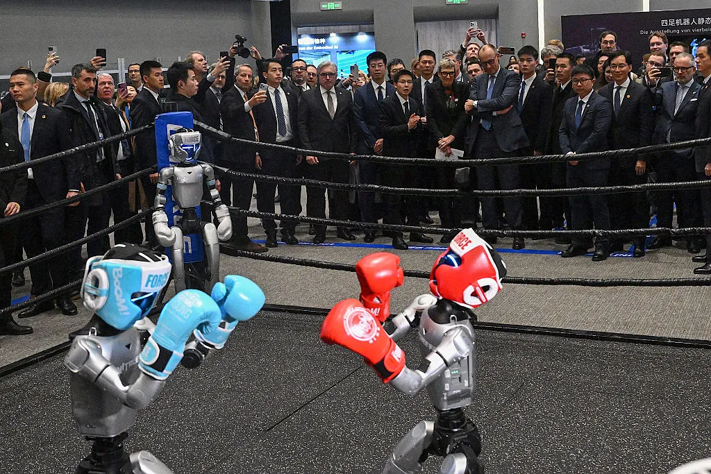 Hard at it. Friedrich Merz (back center R) watches Unitree’s robots boxing in China.