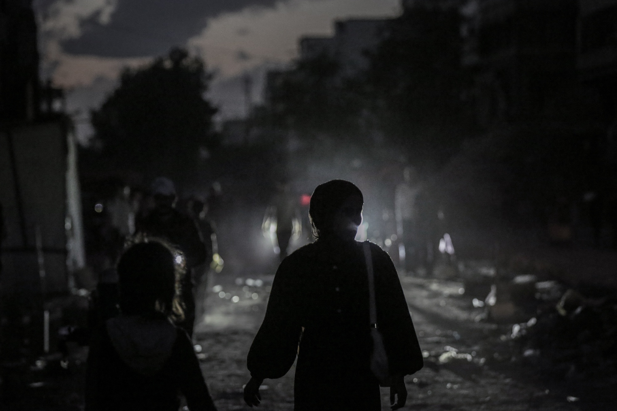 Two figures are backlit by lights from a car as they walk down a street. Other people are visible at the edges of the frame.