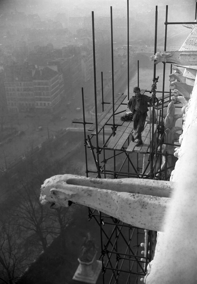 A black and white photo shows a worker standing on scaffolding
