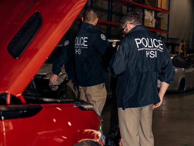 The back of two border police in a warehouse standing beside a vehicle with its hood lifted.