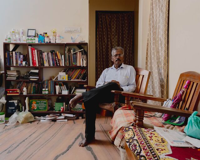 Chinnappan S, a lung cancer patient, sits in a chair by a crowded bookshelf at his home in Chengalpattu, Tamil Nadu. A large stack of medication sits atop the bookshelf.
