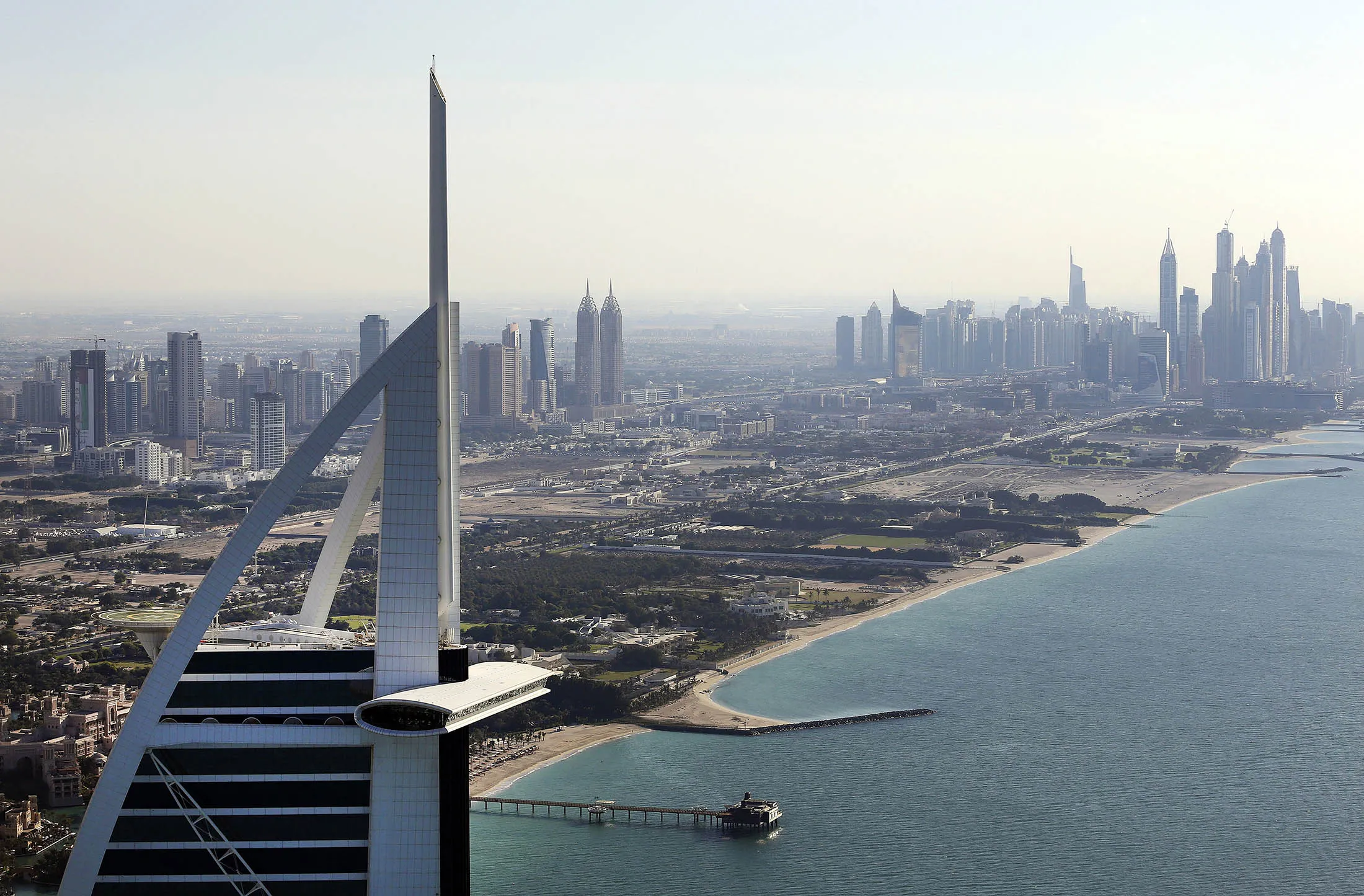 City skyscrapers stand near the coast beyond the tip of the Burj Al Arab luxury hotel.
