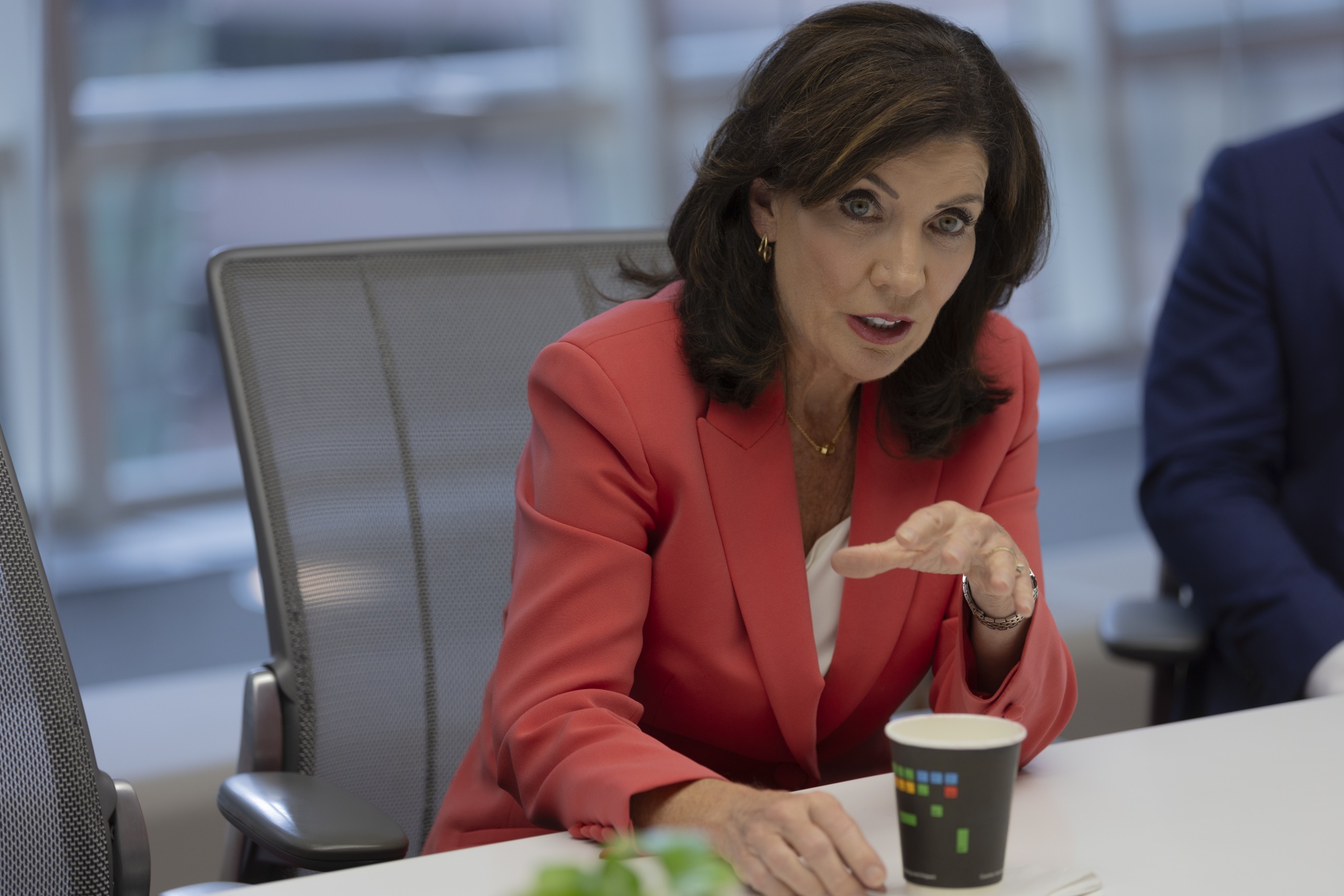 Governor Hochul sits at a conference table wearing a red suit and speaking to someone out of frame.