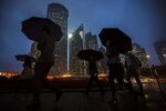 Pedestrians holding umbrellas walk past buildings illuminated at night in the Lujiazui district of Shanghai.
