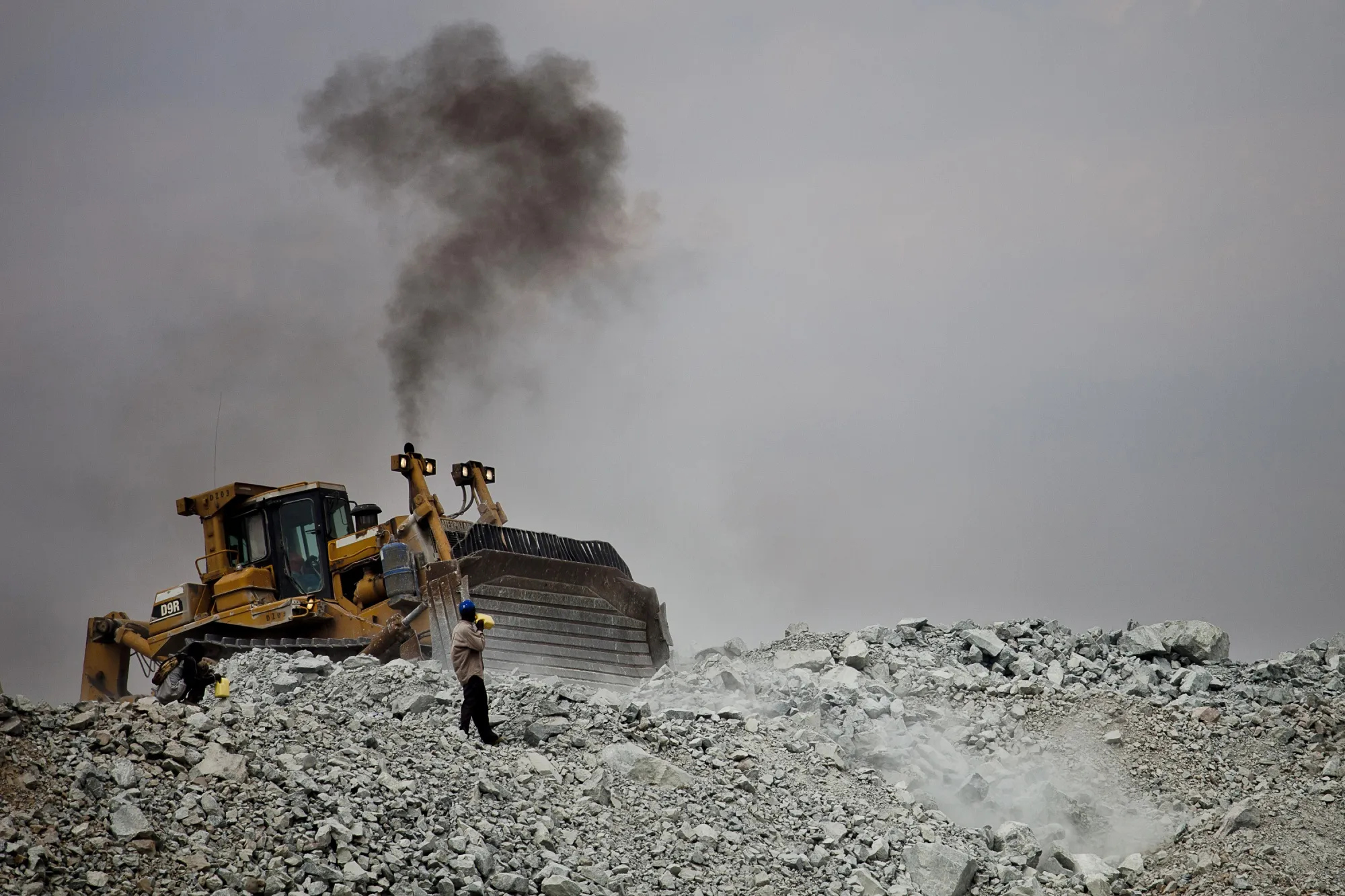 A bulldozer moves rubble as a villager searches for tiny flecks of gold contained in discarded waste rock from Barrick Gold Corp.'s North Mara mine in the district of Nyangoto, Tanzania, on Saturday, July 31, 2010.&nbsp;Skyrocketing bullion prices helped propel&nbsp;Barrick to post its highest quarterly revenue since last year’s&nbsp;merger&nbsp;with Randgold Resources Ltd.