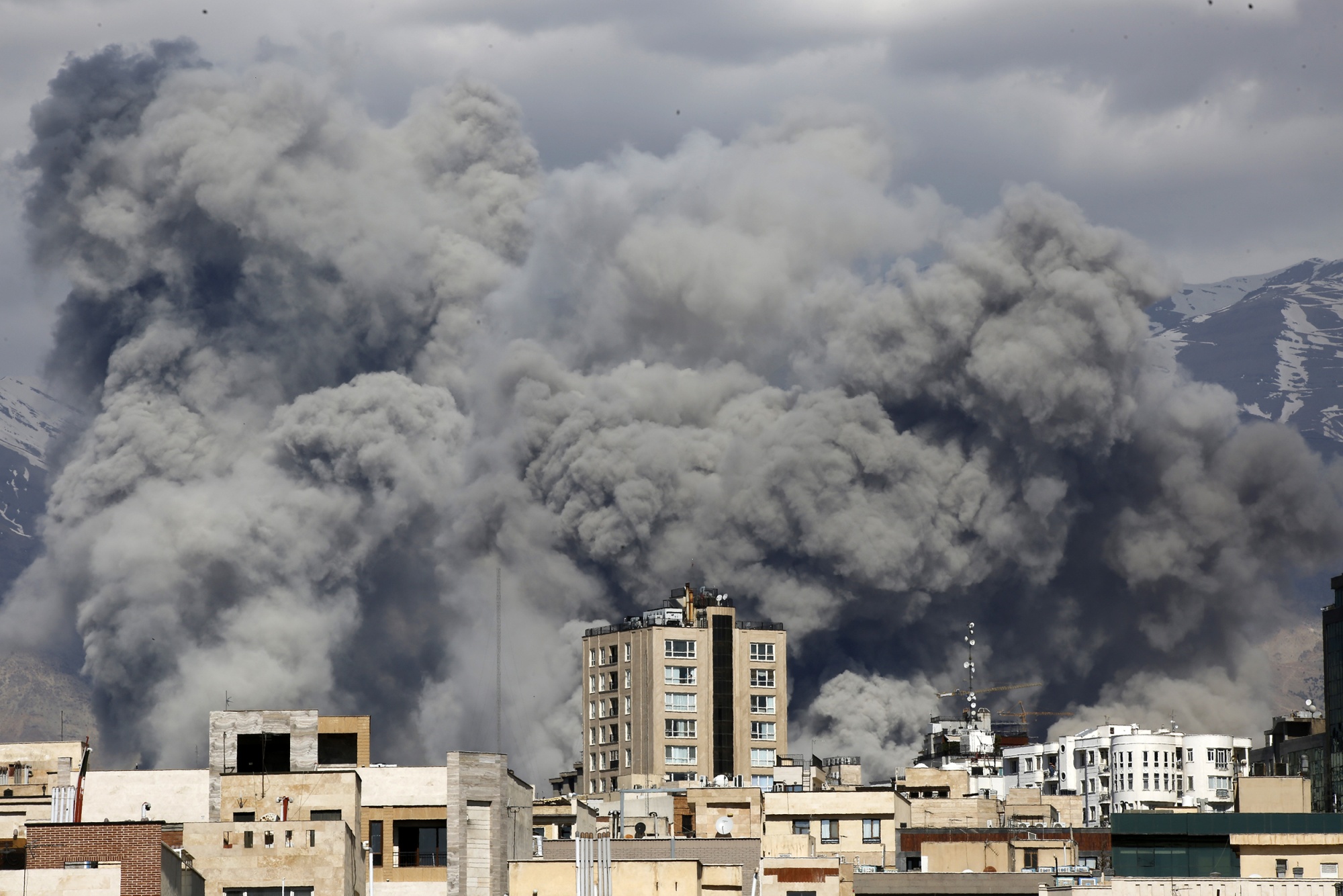 A cloud of smoke rises behind the roofs of buildings. In the background, tall mountains are covered in snow.