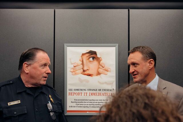 A photo showing head and shoulders of two men chatting, with a "If you see something say something" poster between them. The man on the left is wearing a police uniform and the other is wearing a tan suit.