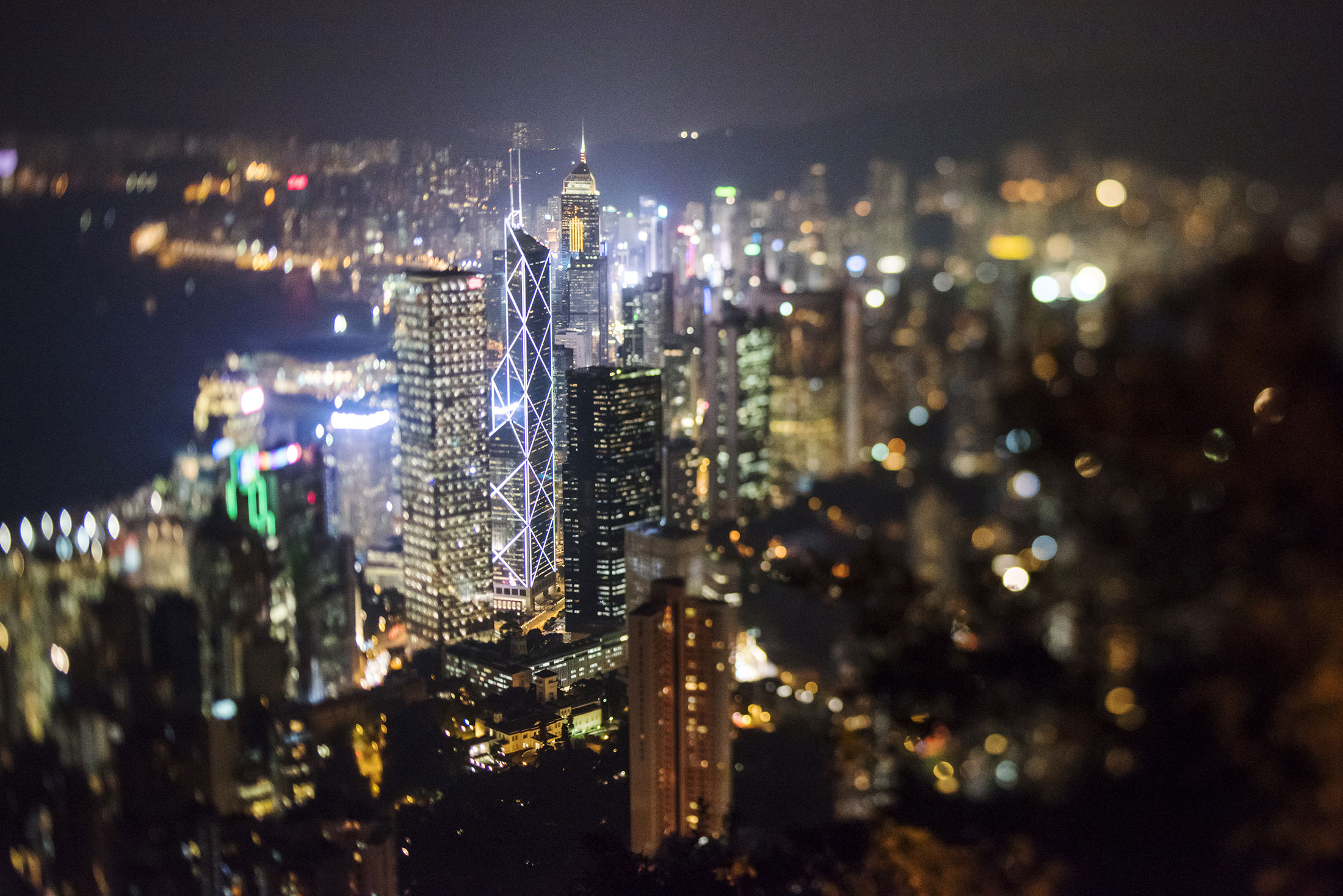 The Bank of China Tower, center left, and other commercial buildings stand illuminated at night in this photograph taken with a tilt-shift lens at night in Hong Kong, China on Friday, Nov. 27, 2015. Secondary private residential property prices dropped 3.9 percent since peaking in September, according to an index published by broker Centaline Property Agency Ltd.
