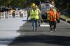 Two road workers walk along an asphalt road that is half treated with a concrete-color surface coating