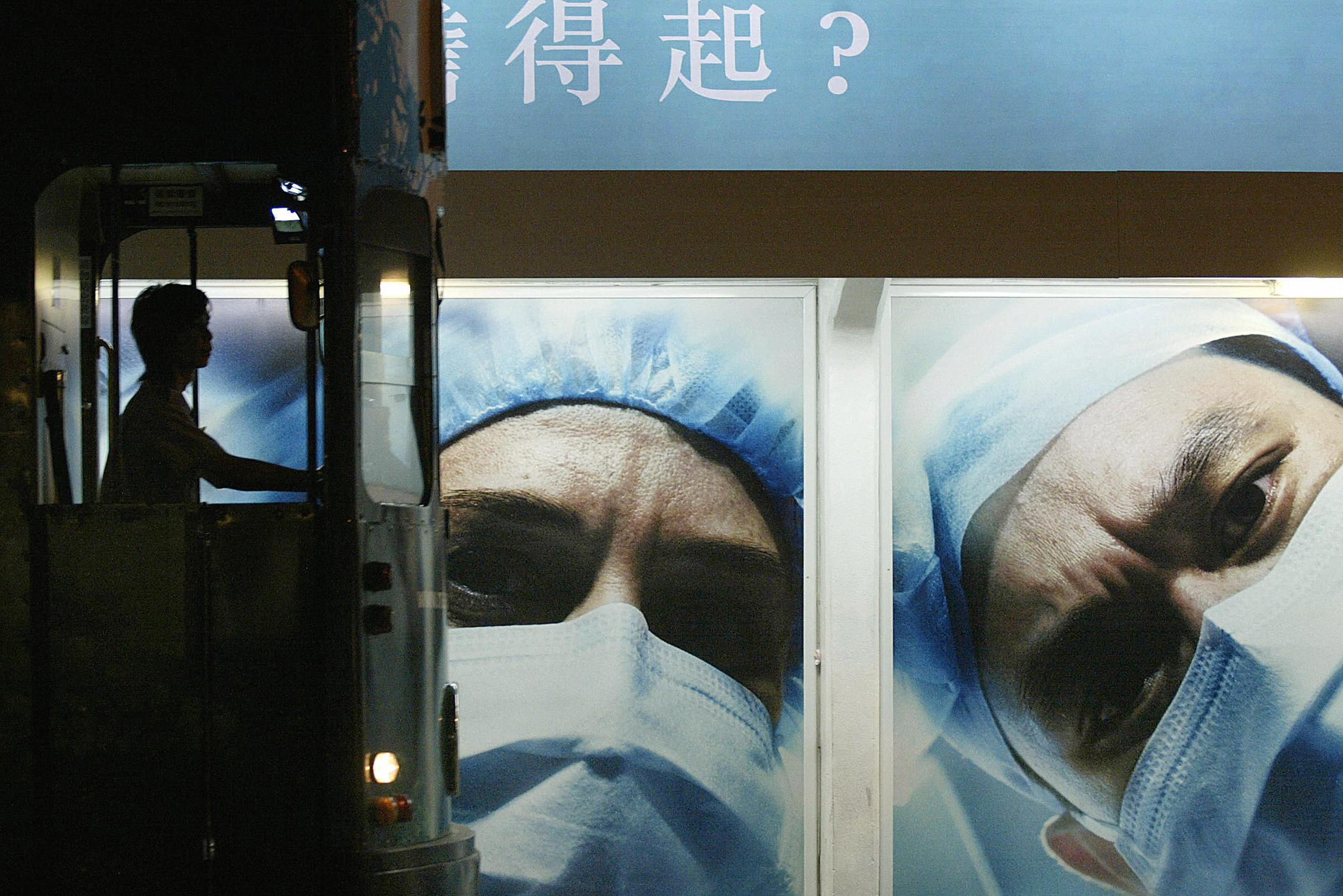 A tram driver is silhouetted at a tram stop in front of a billboard showing medical workers wearing masks in Hong Kong in 2004.