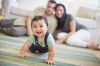 Smiling baby crawling on living room floor with parents watching in the background, out of focus