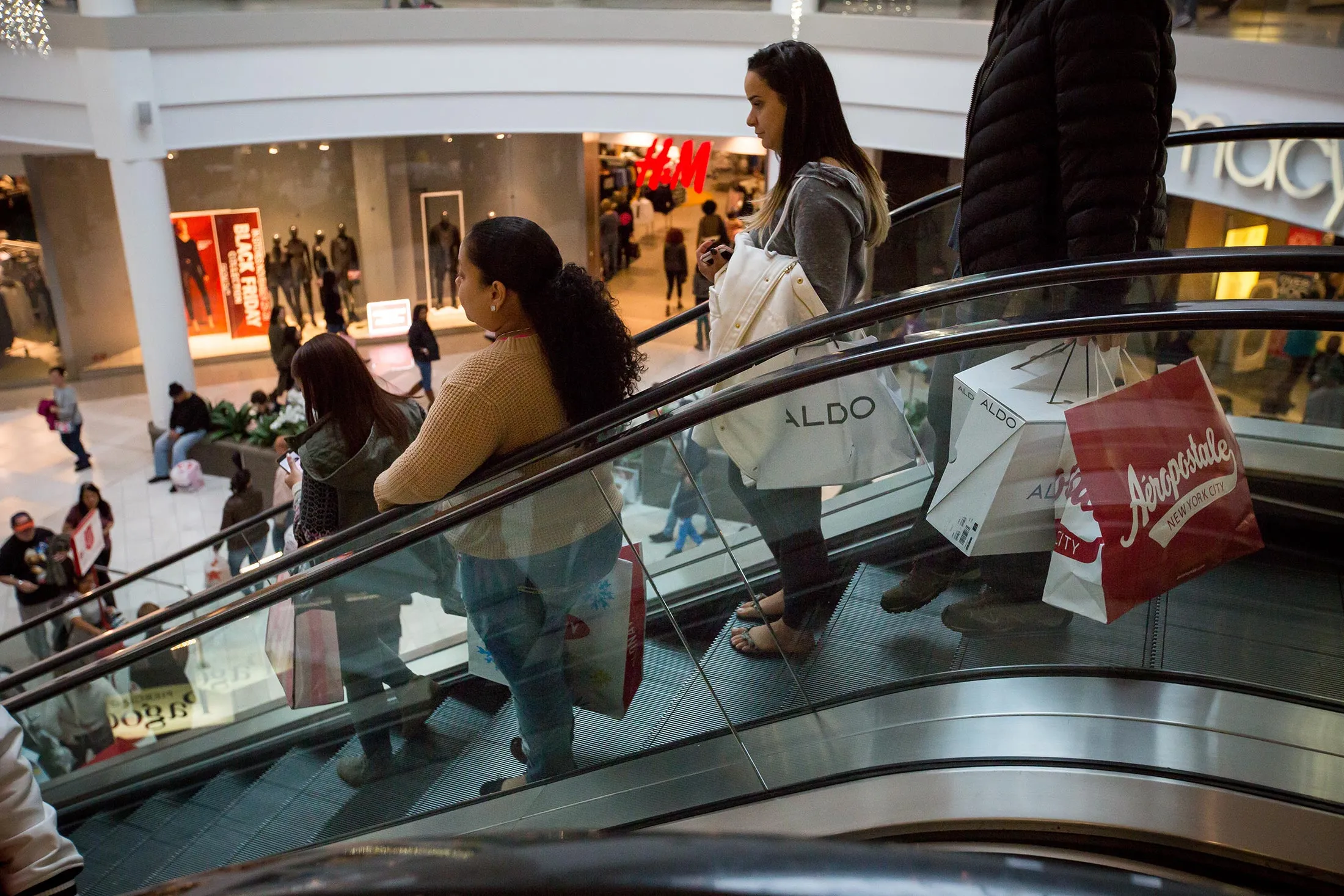 Shoppers ride down the escalator at the Menlo Park Mall in Edison, New Jersey, U.S., on Friday, Nov. 25, 2016. As Black Friday ushers in the year-end shopping rush, chains are touting larger price cuts than in 2015 -- a gamble that maintaining market share is worth squeezing margins.
