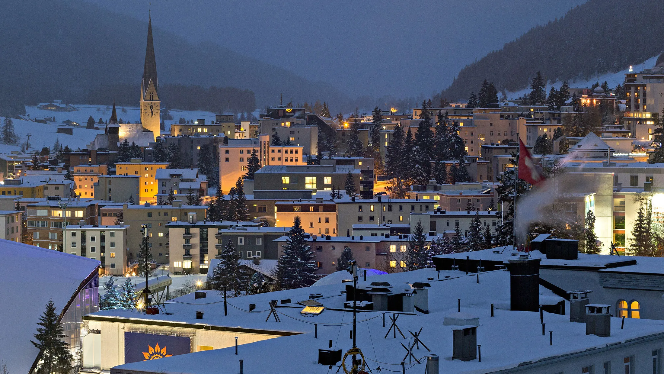 A Swiss national flag flies above a snow covered rooftop as the spire of St. Johann church, left, stands above buildings illuminated at dusk in Davos, Switzerland, on Friday, Jan. 13, 2017. World leaders, influential executives, bankers and policy makers attend the 47th annual meeting of the World Economic Forum (WEF) in Davos from Jan. 17-20.
