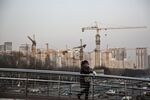 A woman uses a smartphone as she walks a cross a pedestrian footbridge in front of cranes operating at a residential buildings construction site in Beijing, China, on Wednesday, March 2, 2016. Obscured by the focus on the accuracy of China's growth figures is a tumble in estimates for the economy without adjusting for inflation -- a slide that gives a clearer picture of why the country's slowdown has stoked rising concern about its debt burden.
