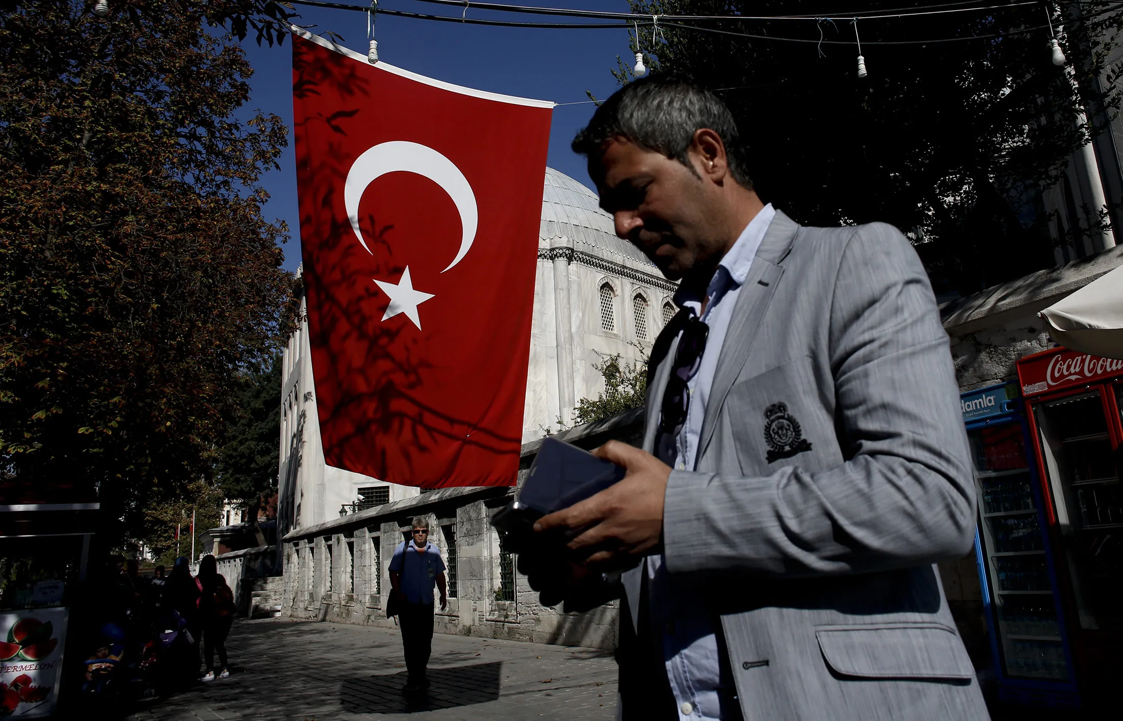 A pedestrian looks at his cellphone as he walks past a Turkish national flag in the Sultanahmet district of Istanbul, Turkey, on Saturday, Oct. 1, 2016. Turkish food prices showed the biggest monthly drop seen in September, bringing annual consumer inflation to the lowest level in over a year and giving more leeway to monetary policy makers to cut borrowing costs this month.
