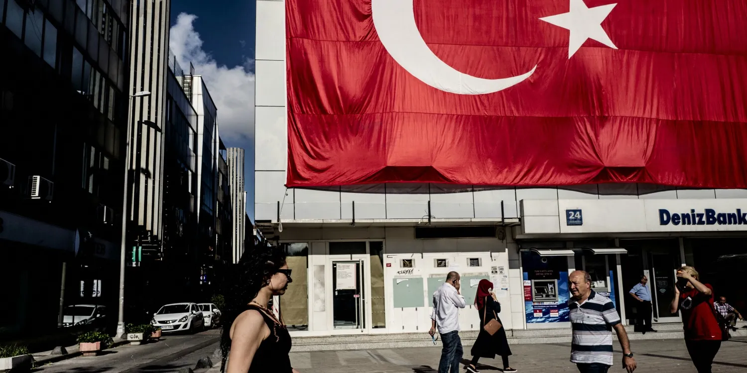 Pedestrians pass a giant Turkish national flag hanging above a DenizBank AS bank branch in Istanbul, Turkey, on Tuesday, July 19, 2016. Turkey's central bank slowed the pace of interest rate cuts at its meeting on Tuesday after the failed coup attempt triggered a sell-off in the currency and sovereign debt.
