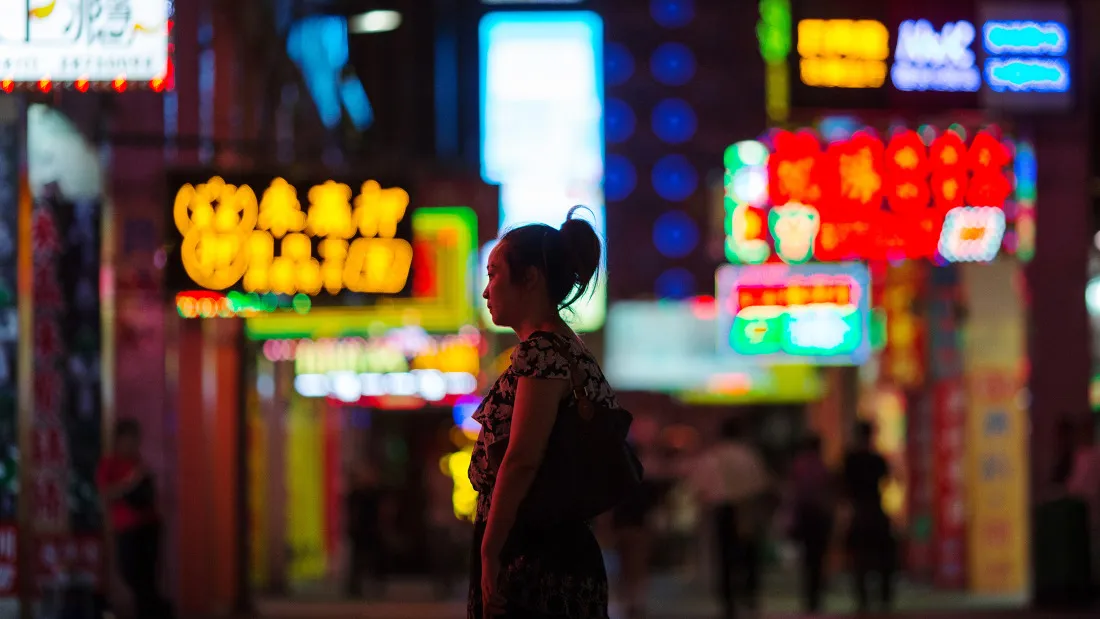 A woman stands in front of neon signs illuminated at night.
