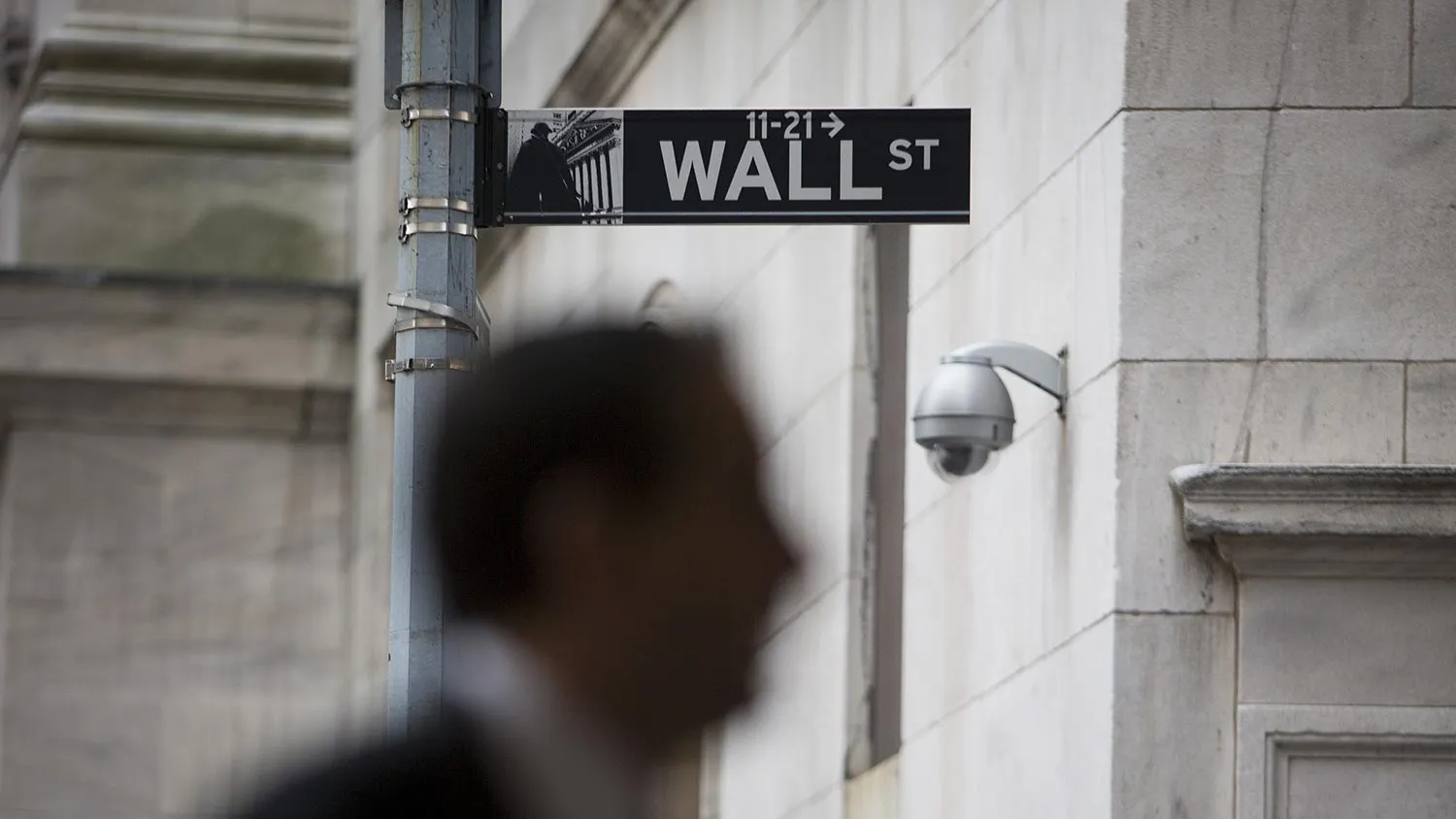 A pedestrian walks past a Wall Street sign and security camera in front of the New York Stock Exchange (NYSE) in New York, U.S., on Wednesday, April 24, 2013.
