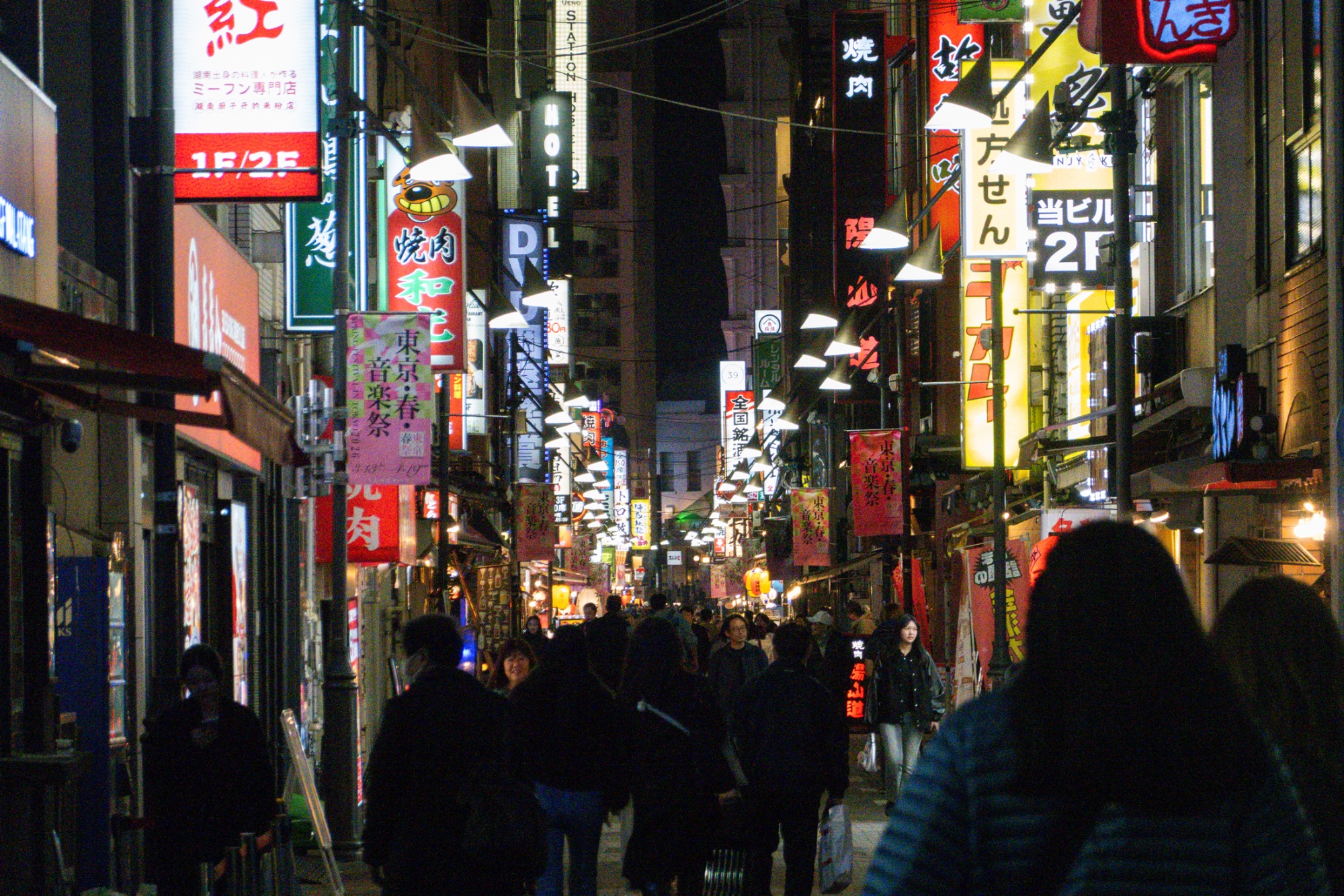 Crowds of pedestrians walk along a narrow street lined with brightly lit restaurants and neon signage in the Ueno district of Tokyo, Japan.