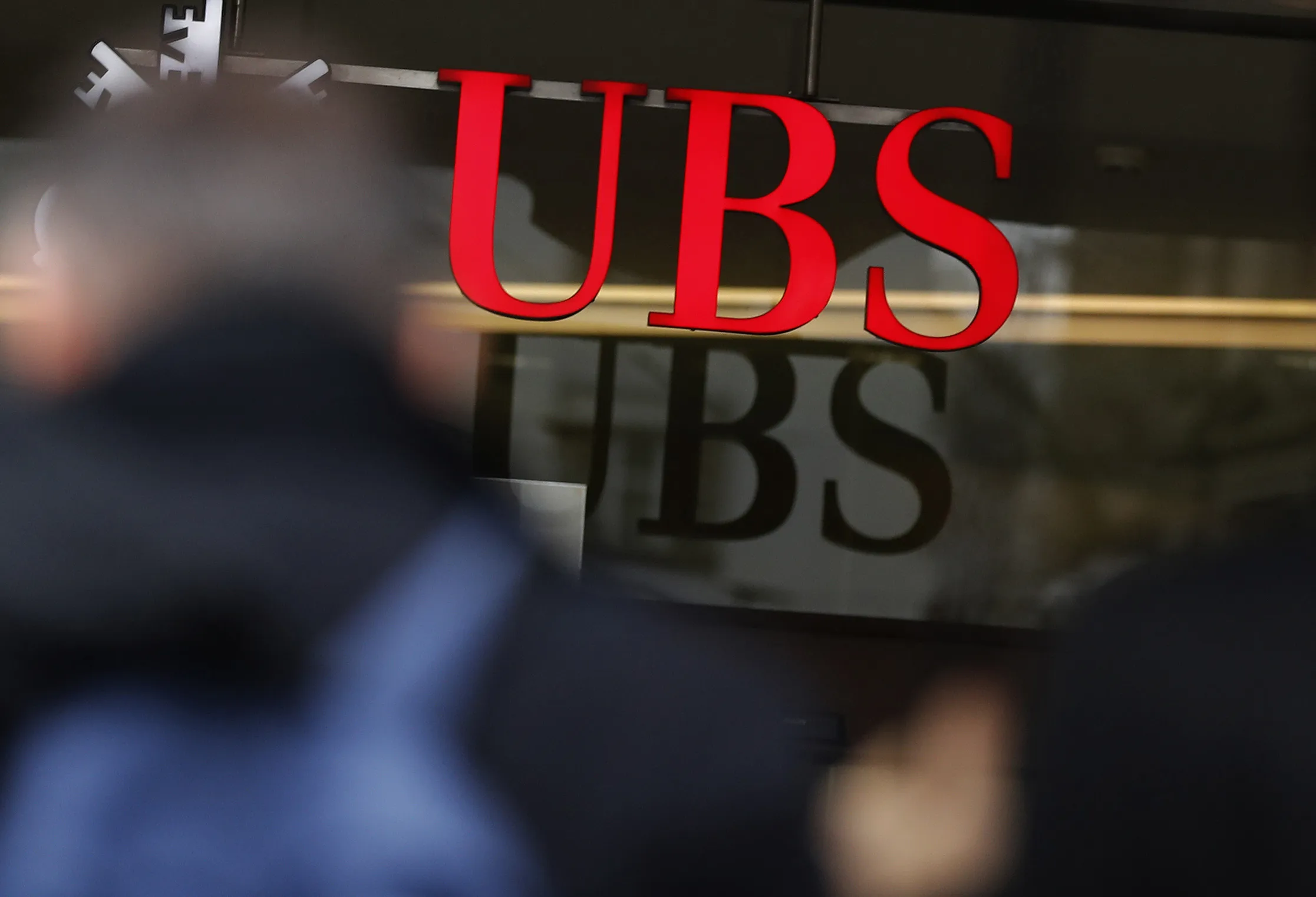 A pedestrian shelters under an umbrella while passing a UBS Group AG bank branch in Zurich.