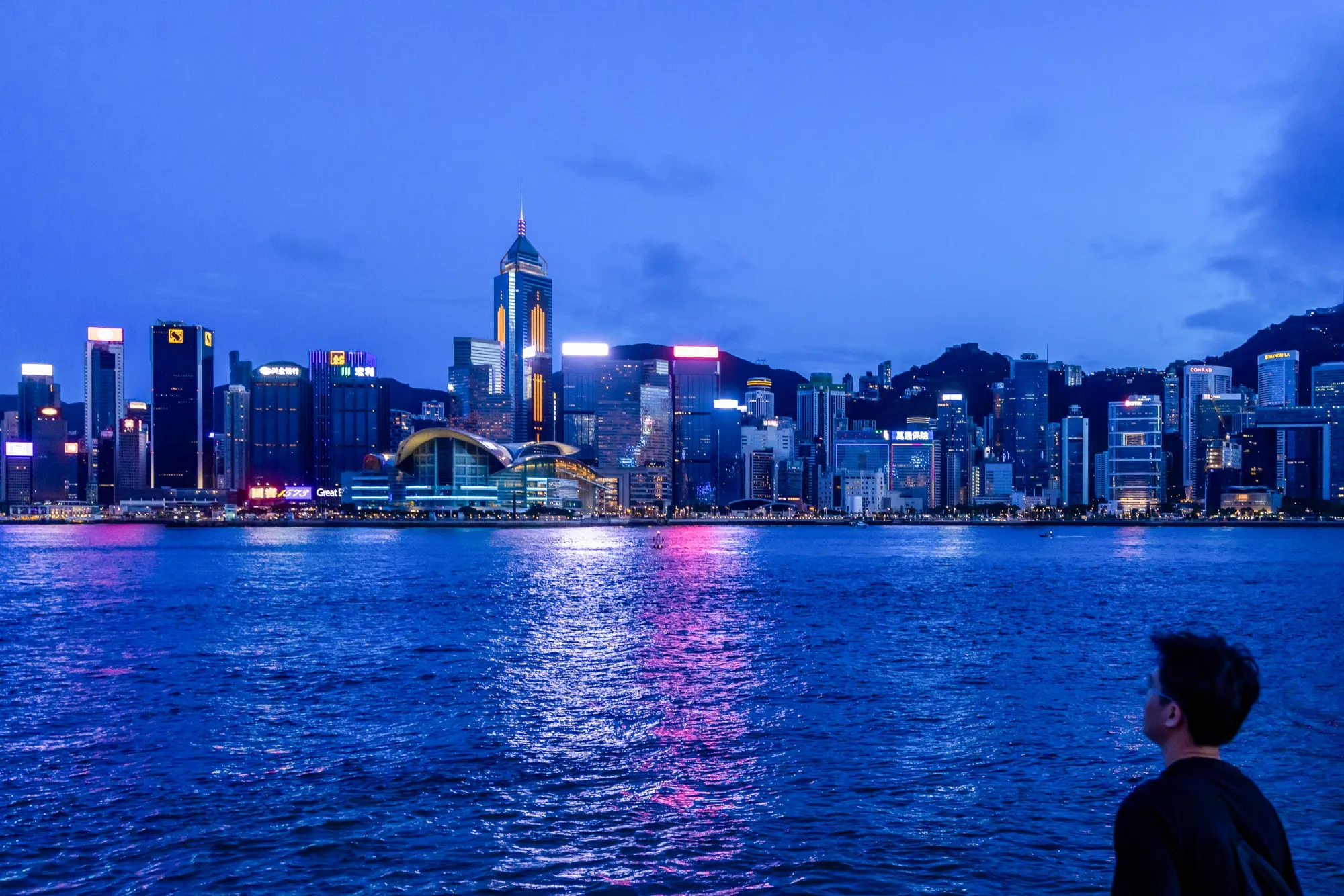 A pedestrian looks out across Victoria Harbour towards the Hong Kong Island skyline in the Tsim Sha Tsui district of Hong Kong, China, on Sunday, June 23, 2024.