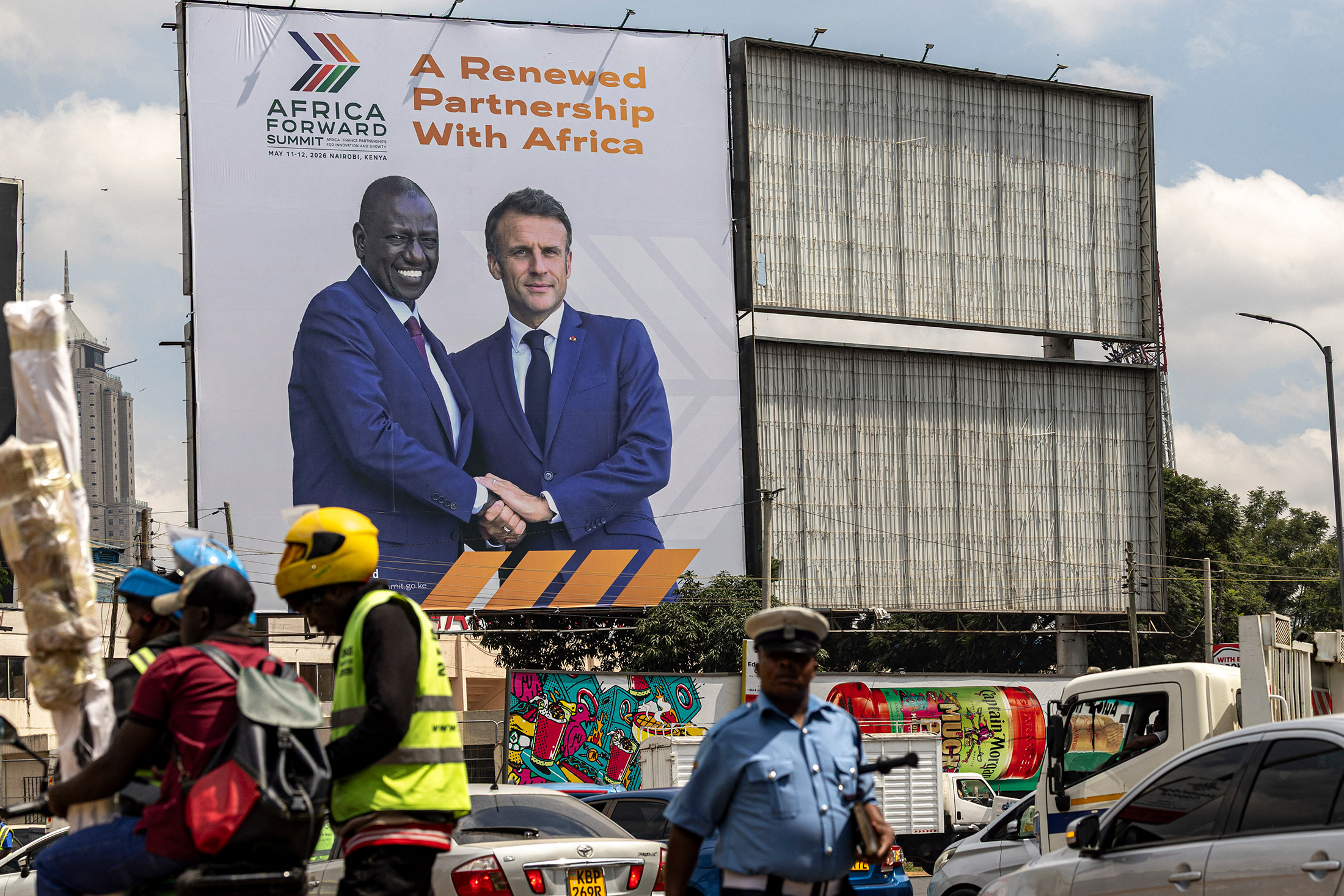 A traffic police officer walks in front of a billboard featuring an image of Kenya's President William Ruto and France's President Emmanuel Macron, promoting the upcoming joint Kenya-France "Africa Forward Summit" in Nairobi on May 6, 2026. Photographer: SIMON MAINA /AFP/Getty Images