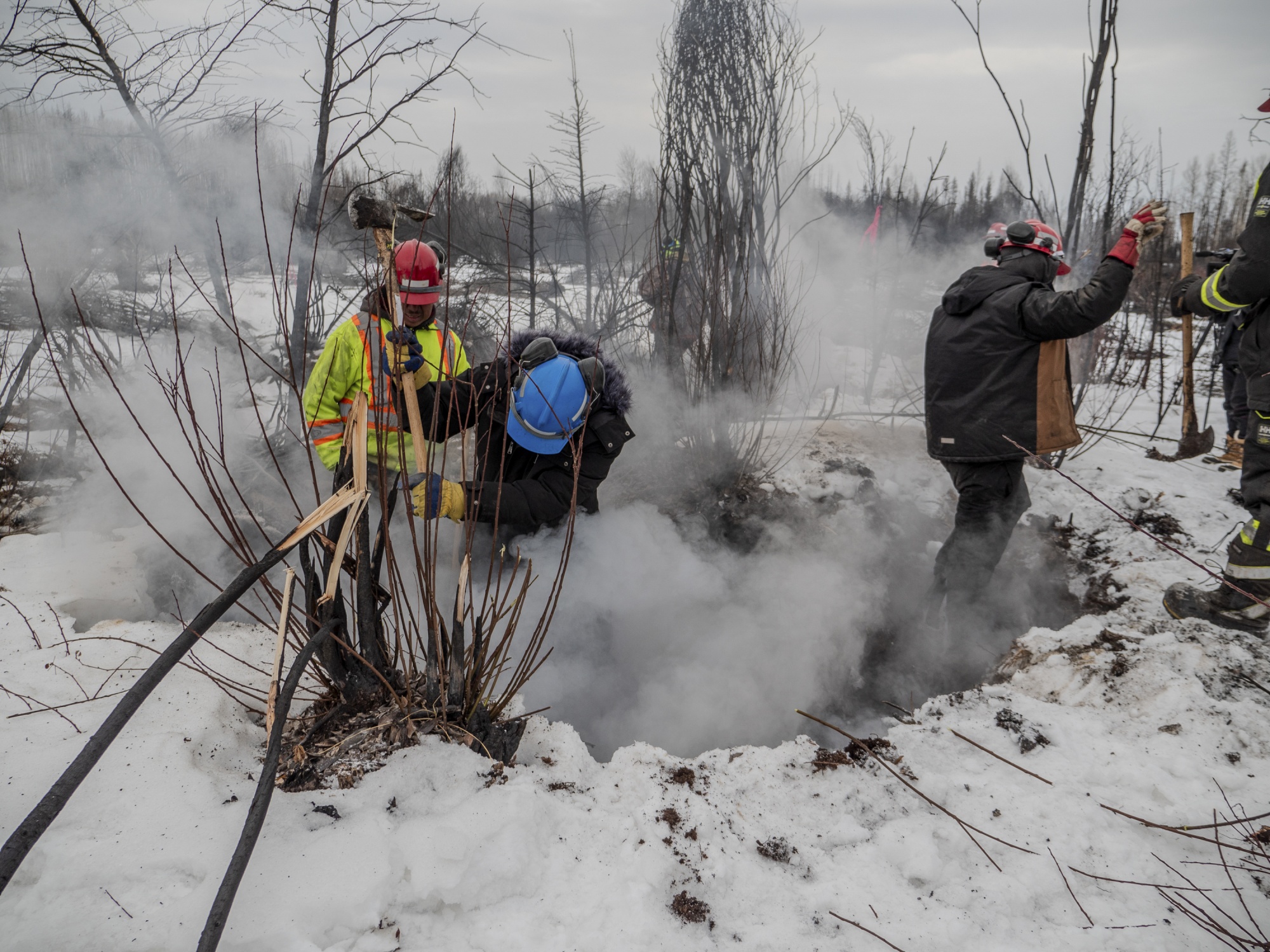 Three firefighters shown waist-deep in snow-topped peatland. They are in winter gear with axes in hand, responding to an underground fire where smoke is billowing out of the ditch they are standing in.